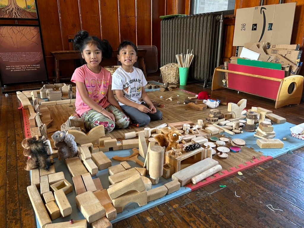 Two children sit beside a large wooden toy city built from blocks and cardboard in a room.