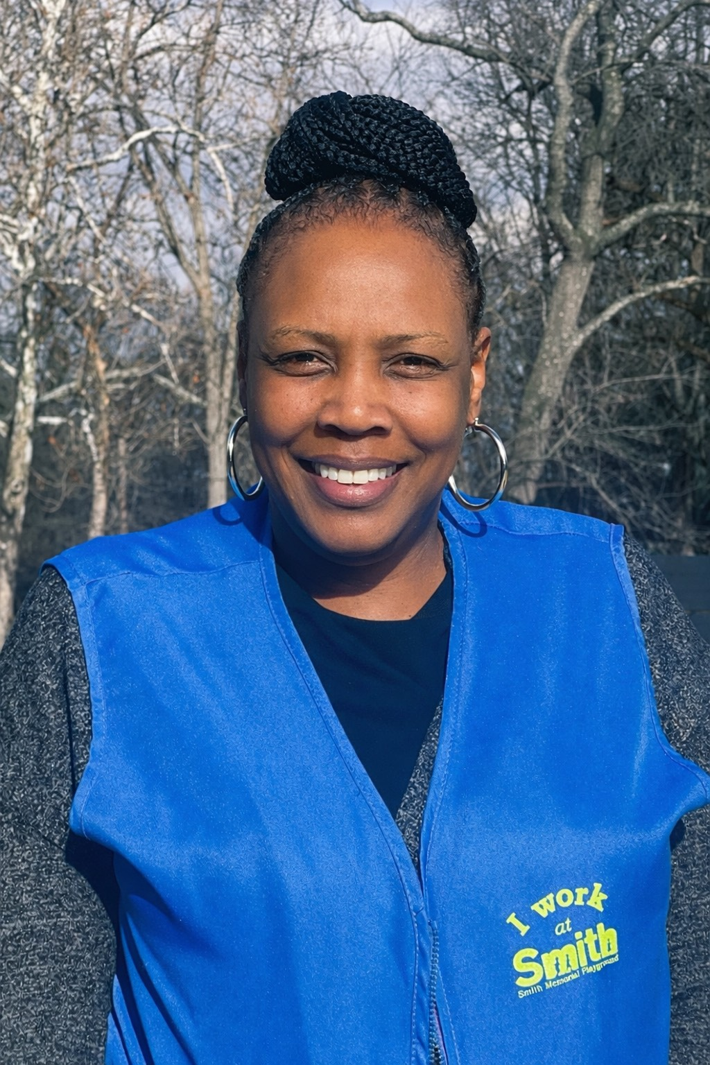 Smiling woman in a blue vest with yellow text, standing outdoors among leafless trees