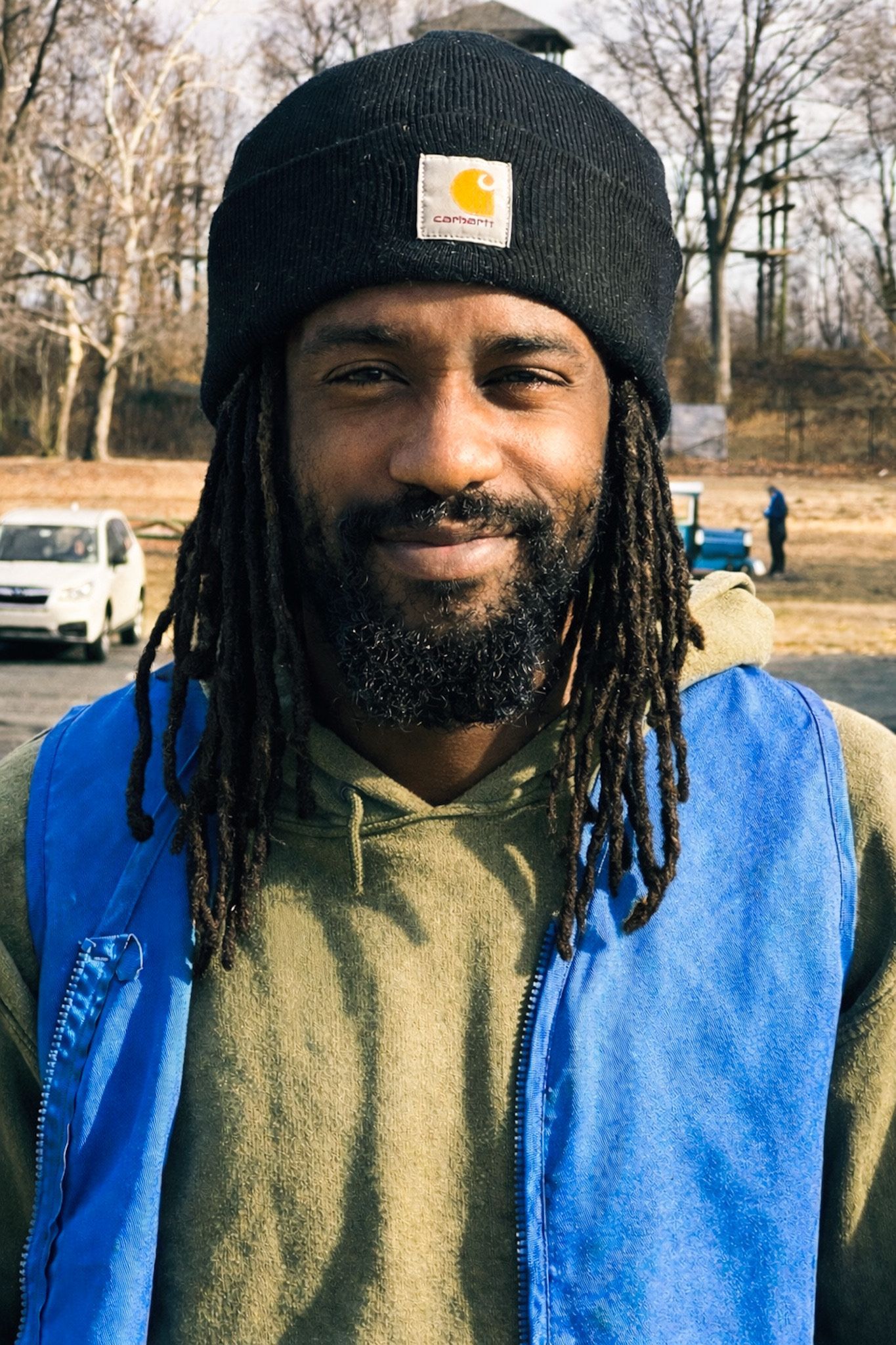 Smiling man in a black knit hat and blue vest standing outdoors in a wooded area