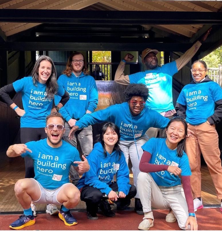 Group of smiling people in blue shirts posing together outdoors under a wooden shelter