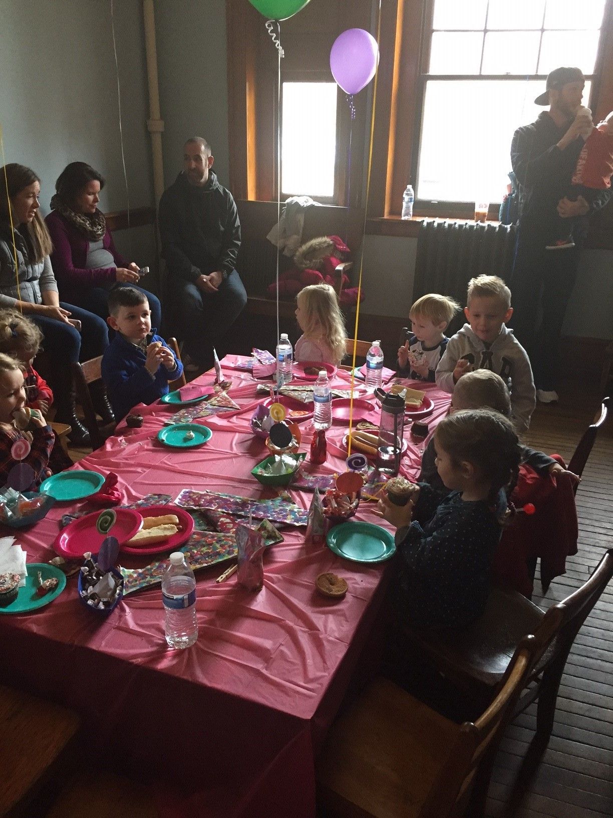 Children seated around a pink birthday table with plates, balloons, and cake in a dim room