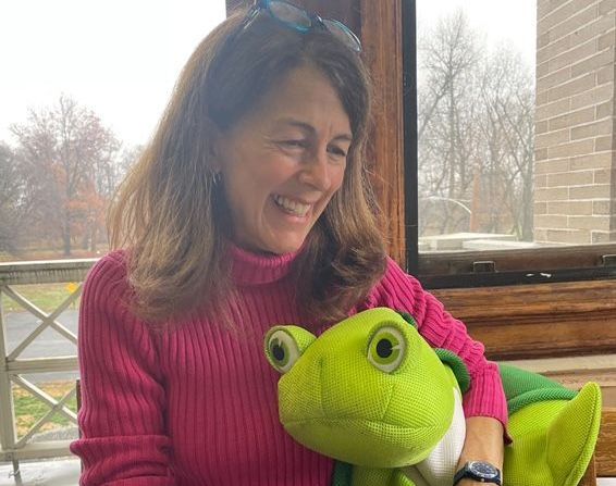 Woman smiling beside a bright green puppet on a porch, with trees and a brick wall in the background.