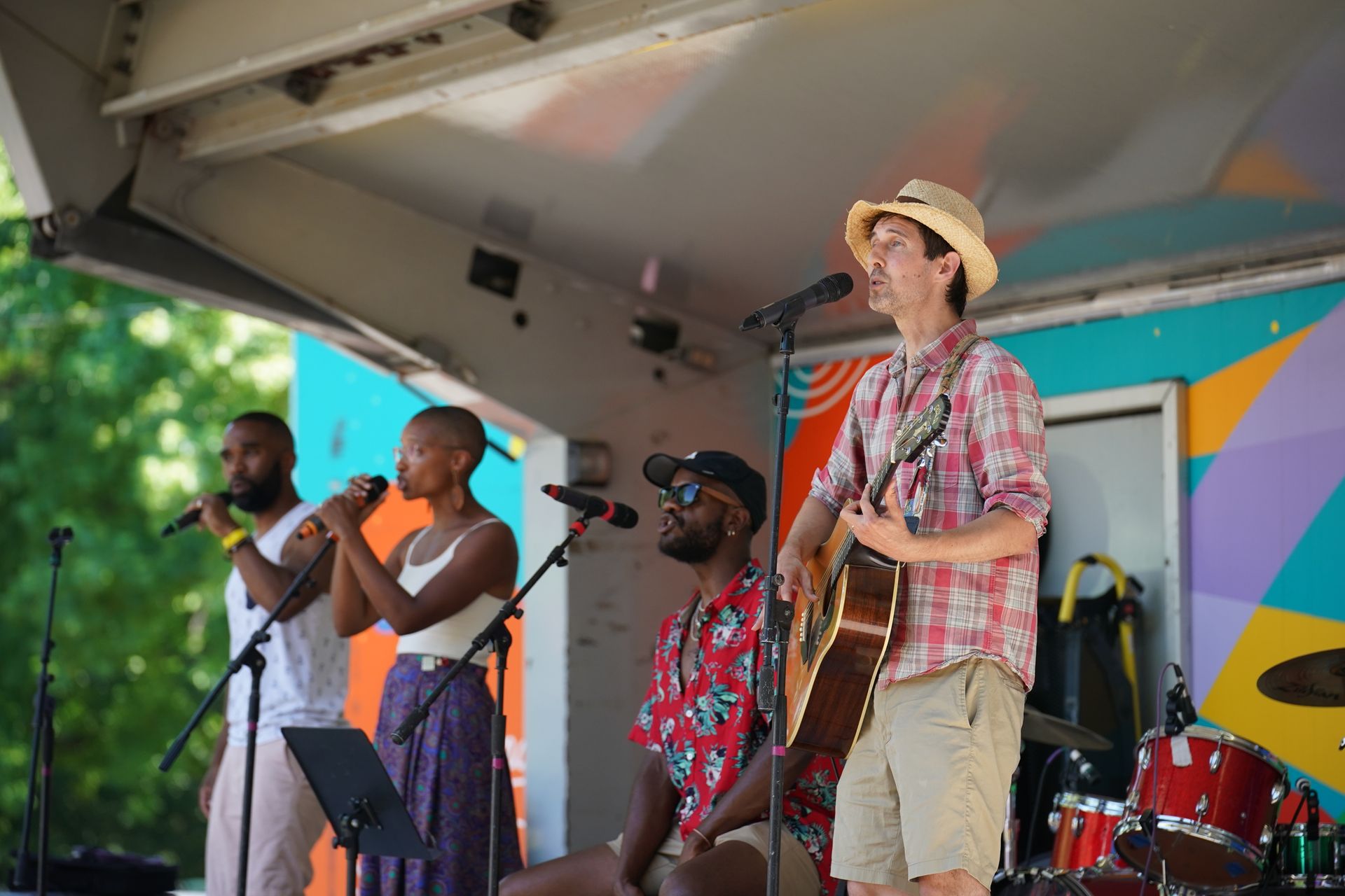Band performing on an outdoor stage, with singer, guitarist, and drummer under a canopy.