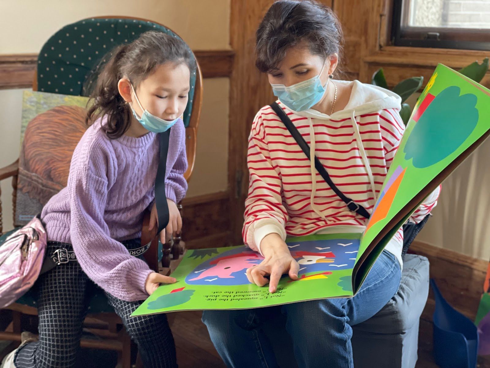 Two children wearing masks read a colorful pop-up book together on a couch.
