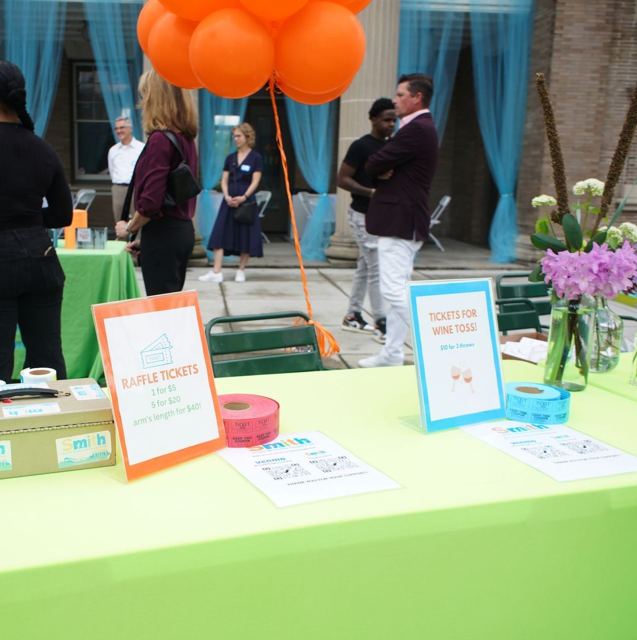 Outdoor event booth with lime-green table, orange balloons, and a few people mingling in the background