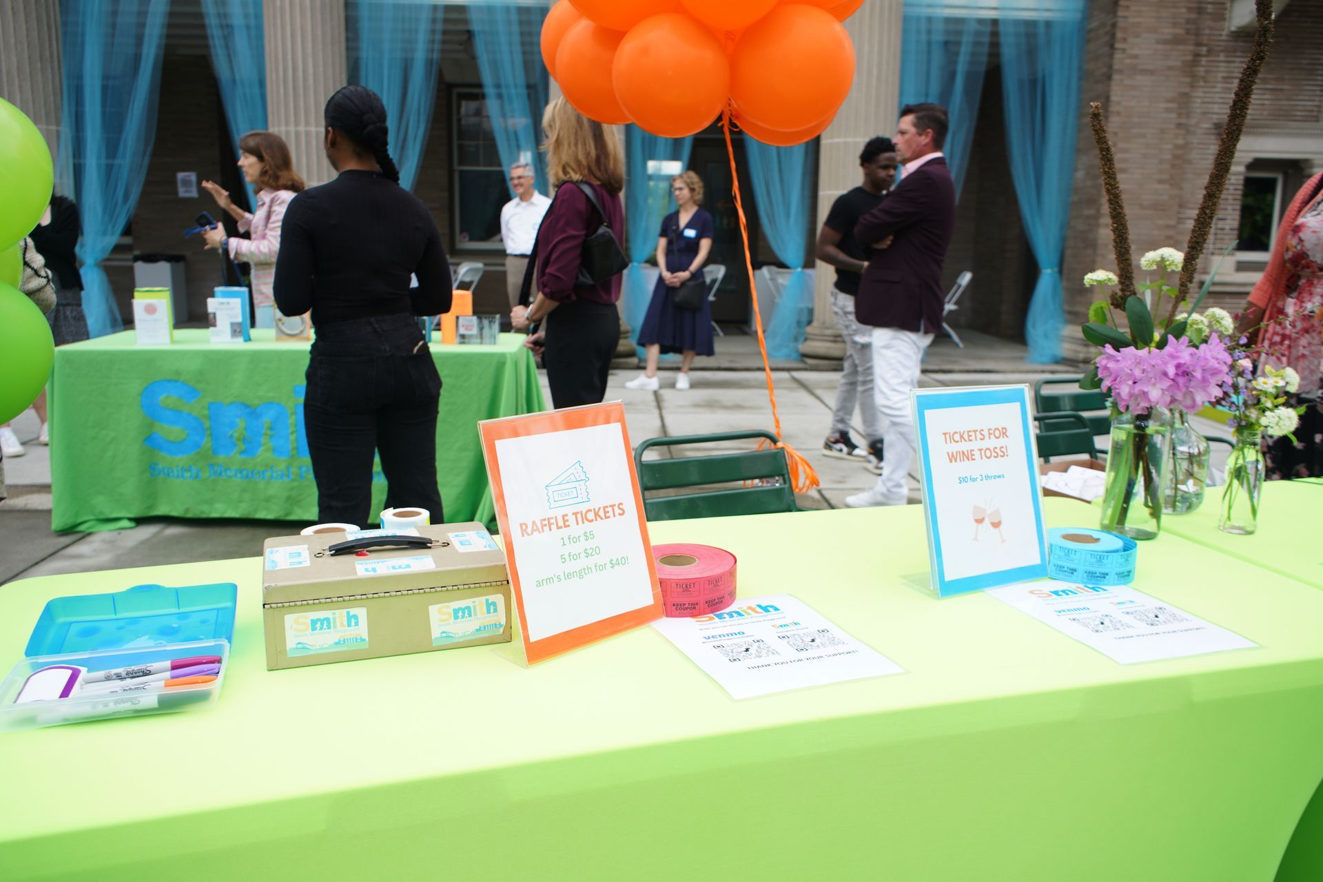 Outdoor booth with lime-green tablecloth, balloons, posters, and people chatting in the background.