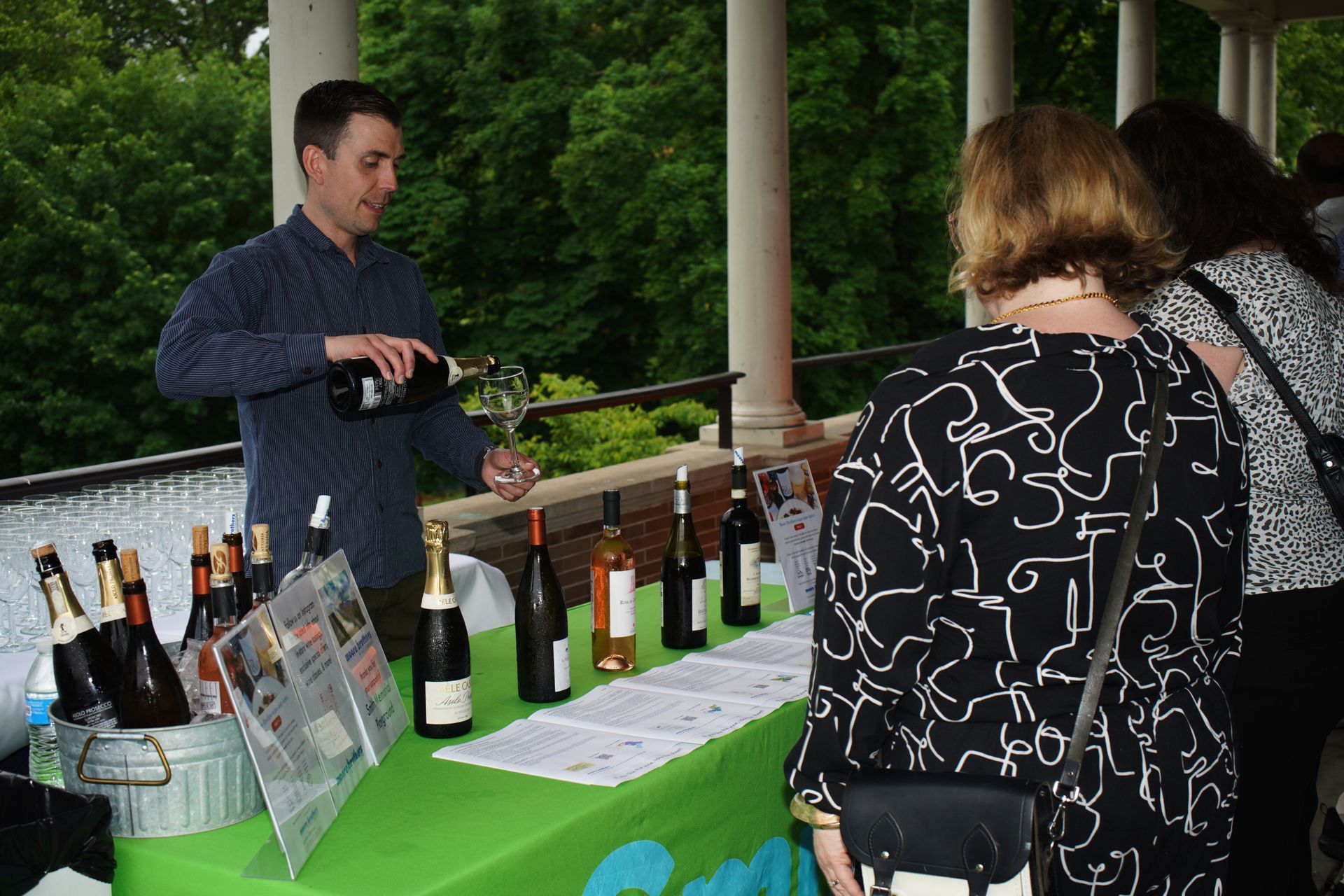 Man pouring wine samples at an outdoor tasting table with bottles and visitors nearby