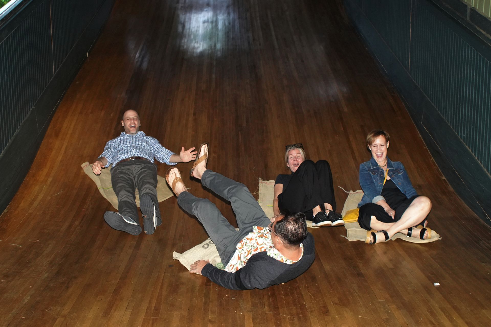 Four people sitting and lying on a wooden bowling lane inside a dim alley