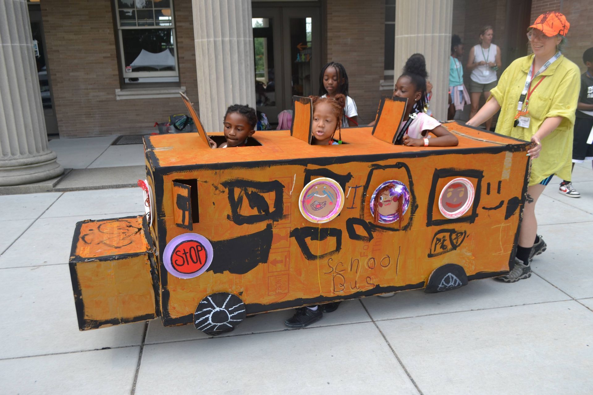 Children ride in an orange cardboard train costume during an outdoor event.