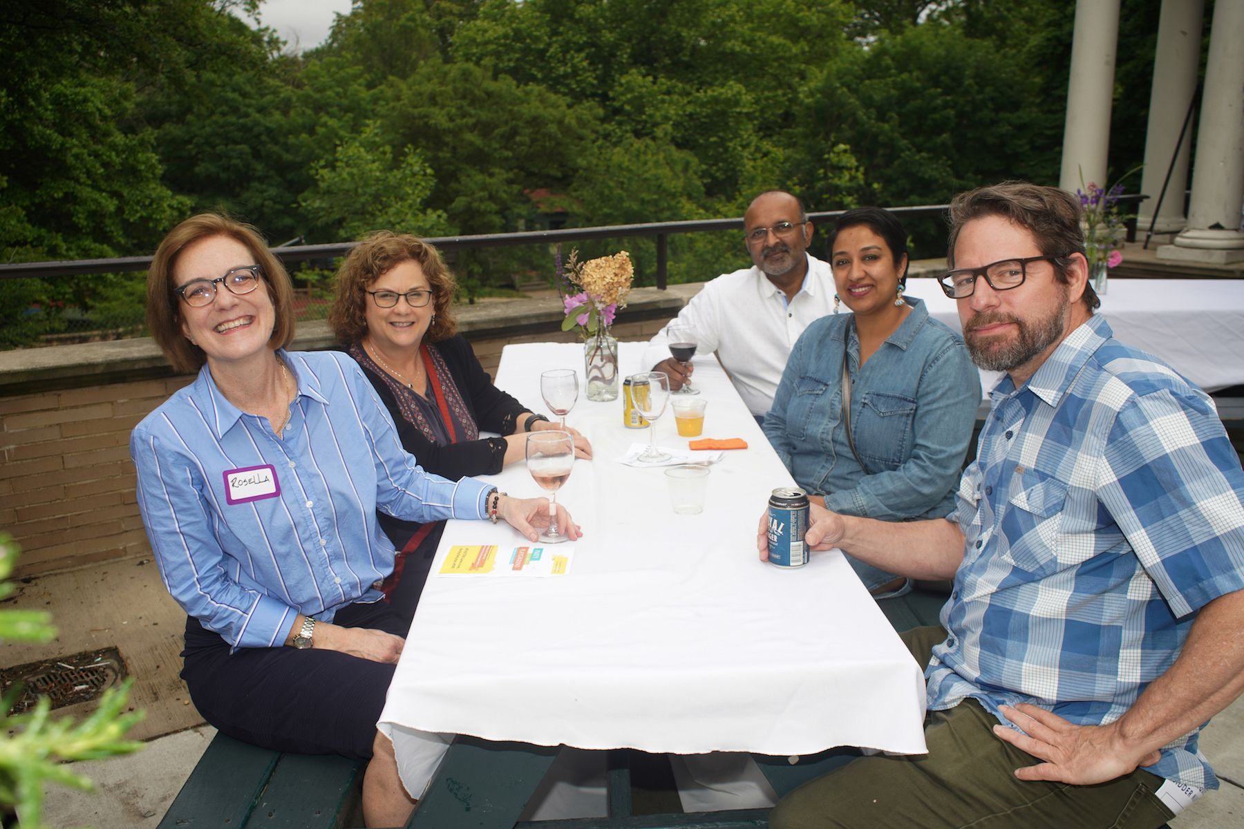 Five people smiling around a picnic table outdoors with drinks and snacks, surrounded by trees