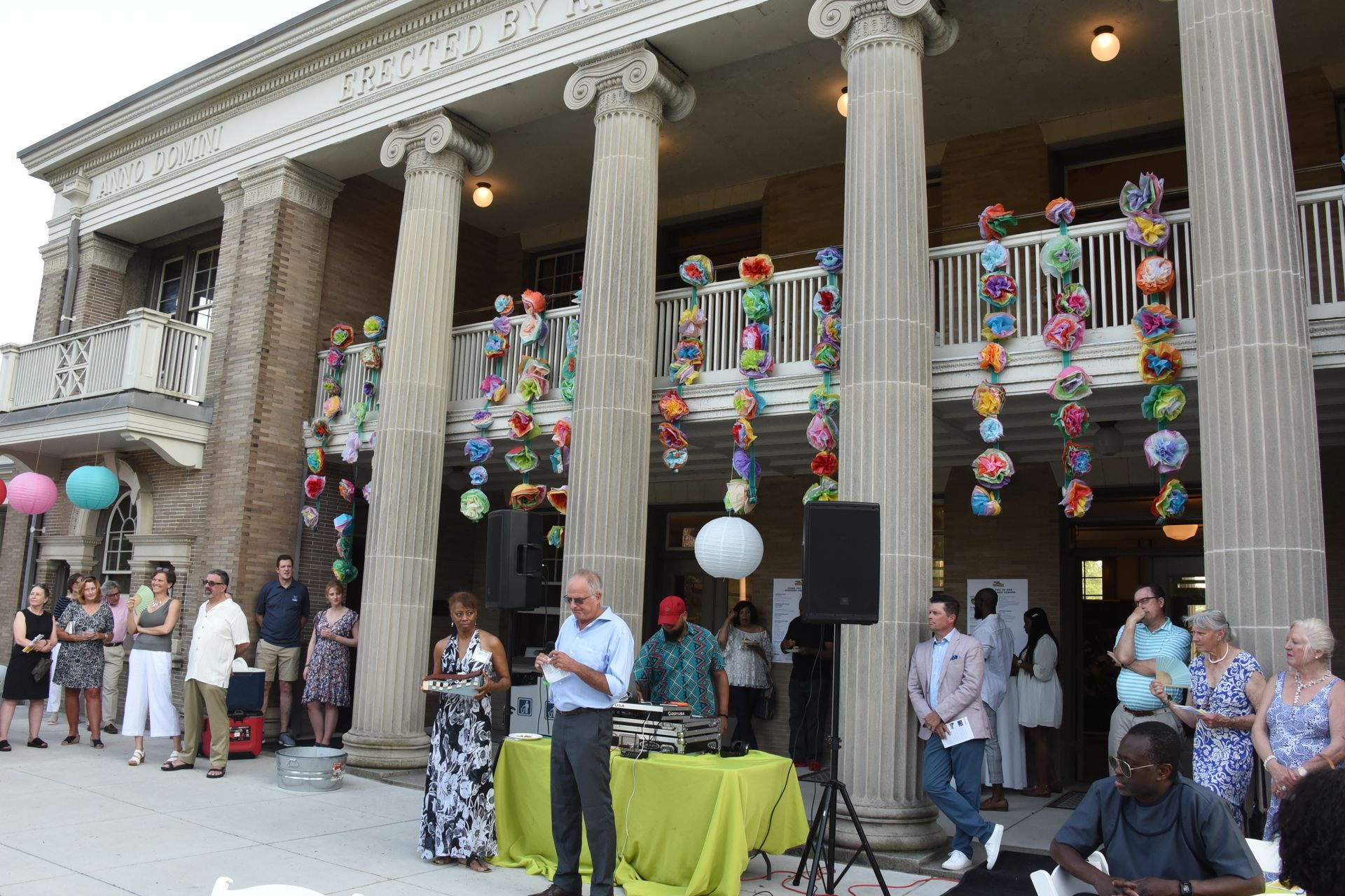 People gathered on a decorated building’s steps for an outdoor celebration, with balloons and speakers.