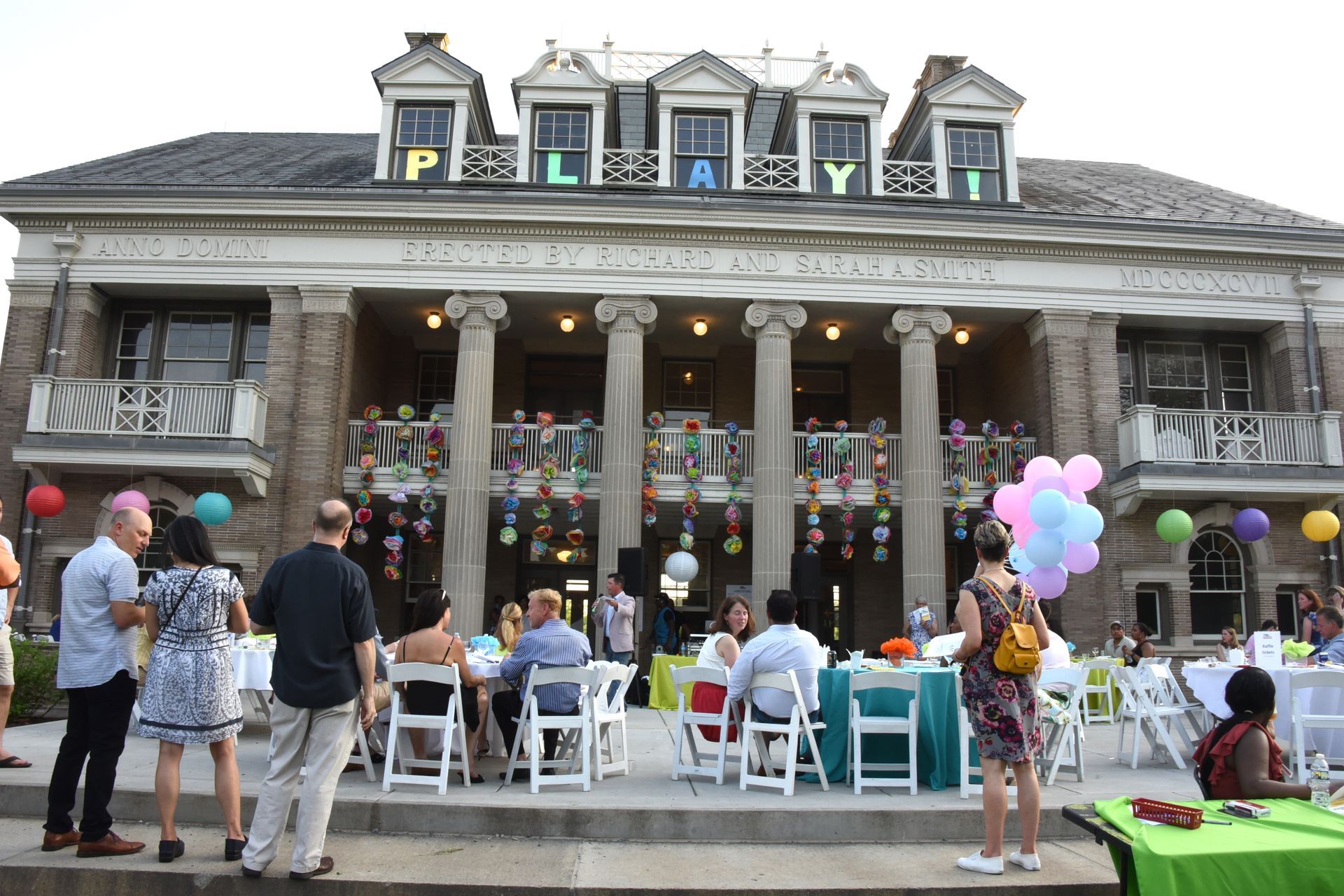 Outdoor gathering at a gray historic building with people seated under a porch and pastel balloons nearby