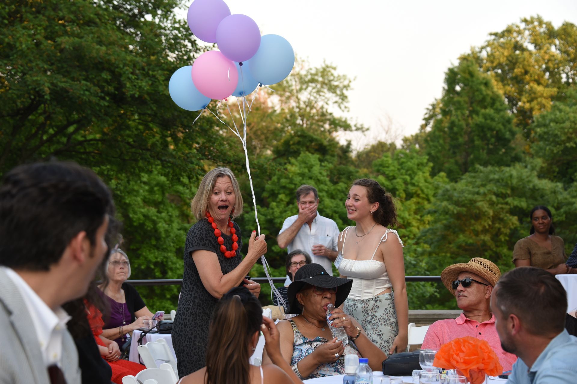 Outdoor party with people gathered around a woman holding pastel balloons and speaking to guests