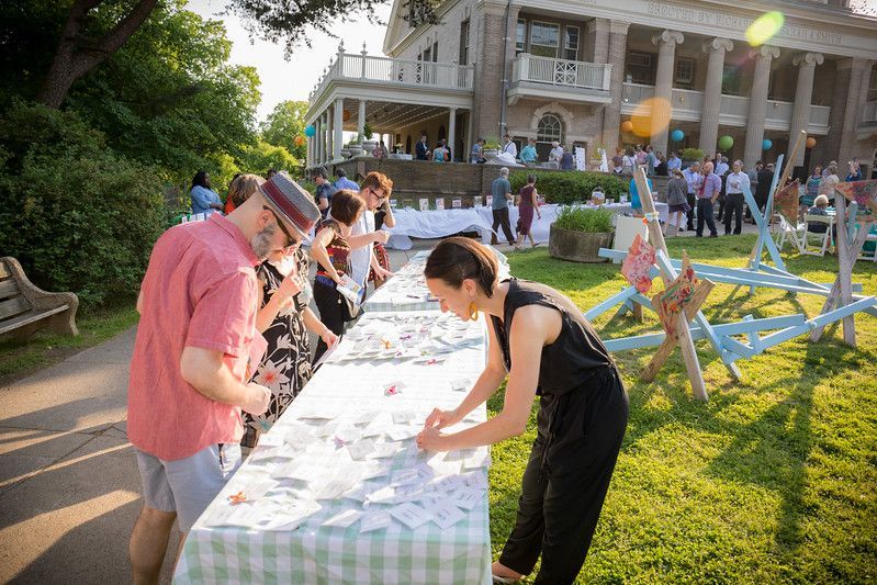 People signing a long table at an outdoor gathering on a lawn by a large house