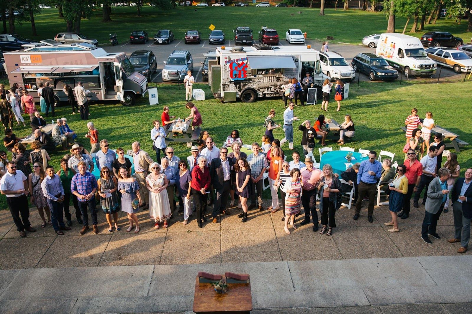 Crowd gathered outdoors at a park, with food trucks and parked cars in the background