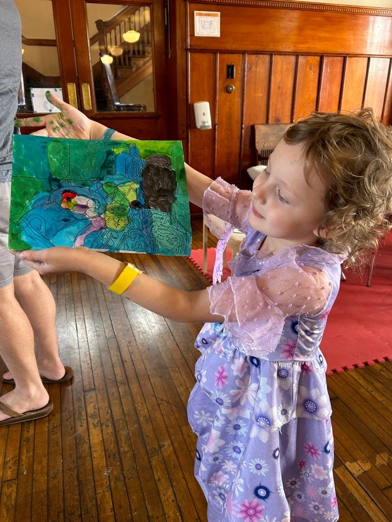 Child in a purple dress holding up a colorful painting indoors on a wooden floor