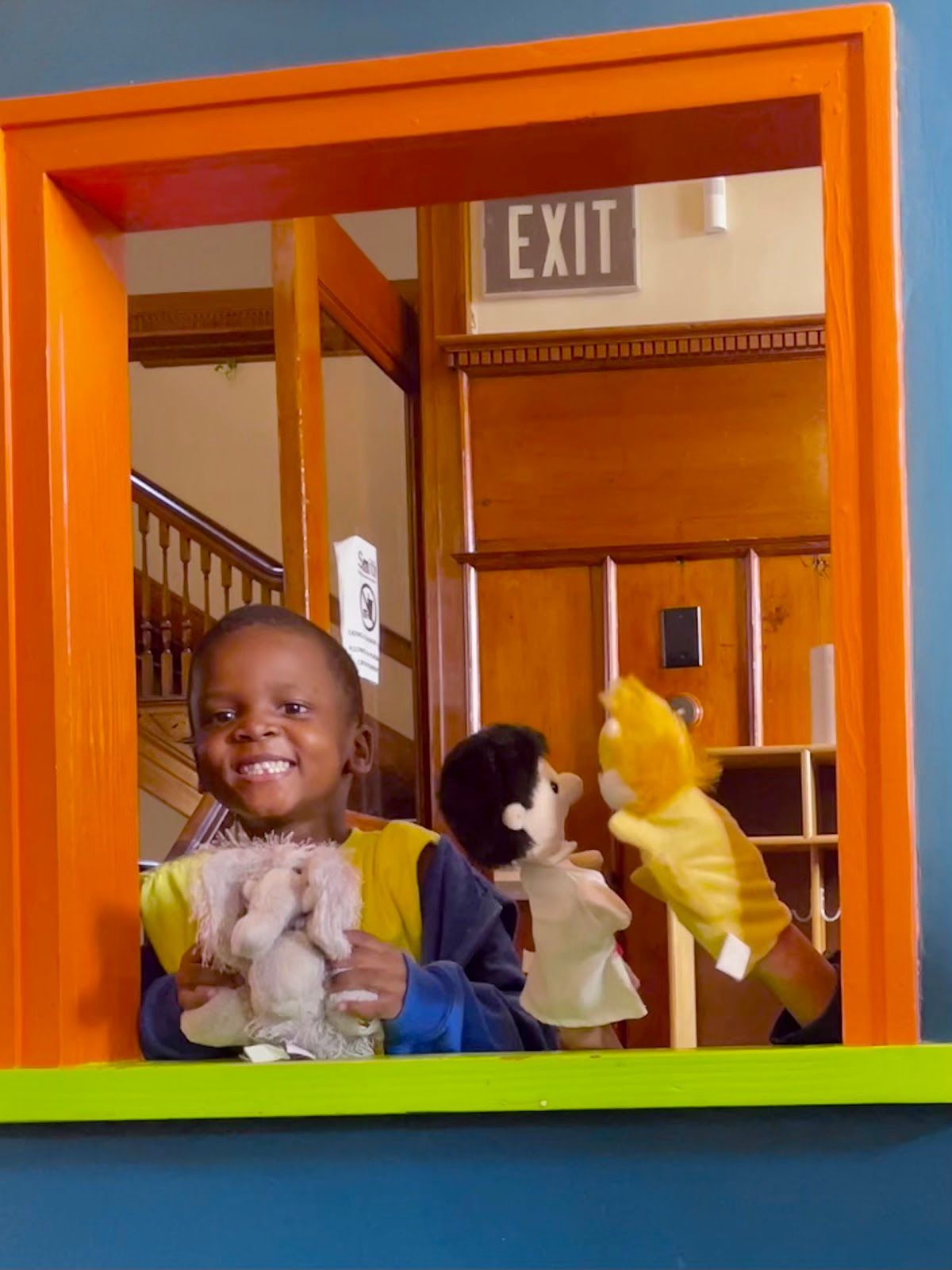 Child smiling behind a play window, holding puppets in a bright orange indoor play area.