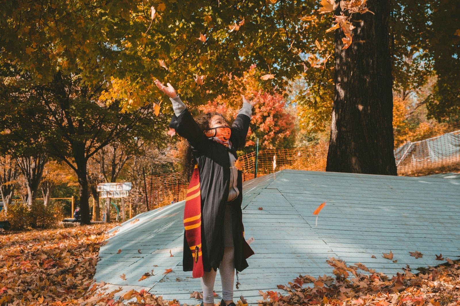 Person in a dark coat with arms raised among autumn trees and fallen leaves near a white dome.