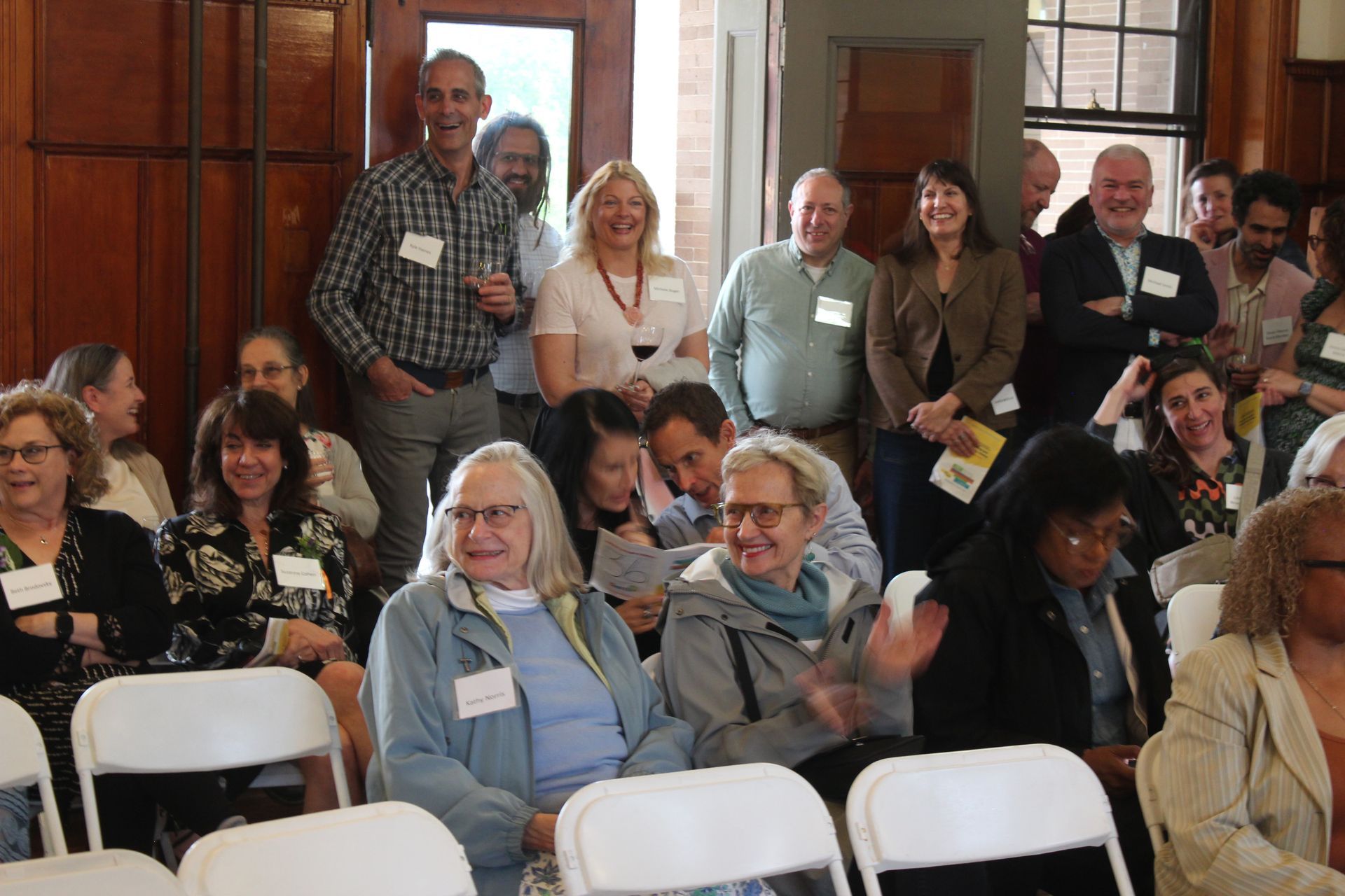 Audience seated in a meeting room, with several people standing and smiling at the back.