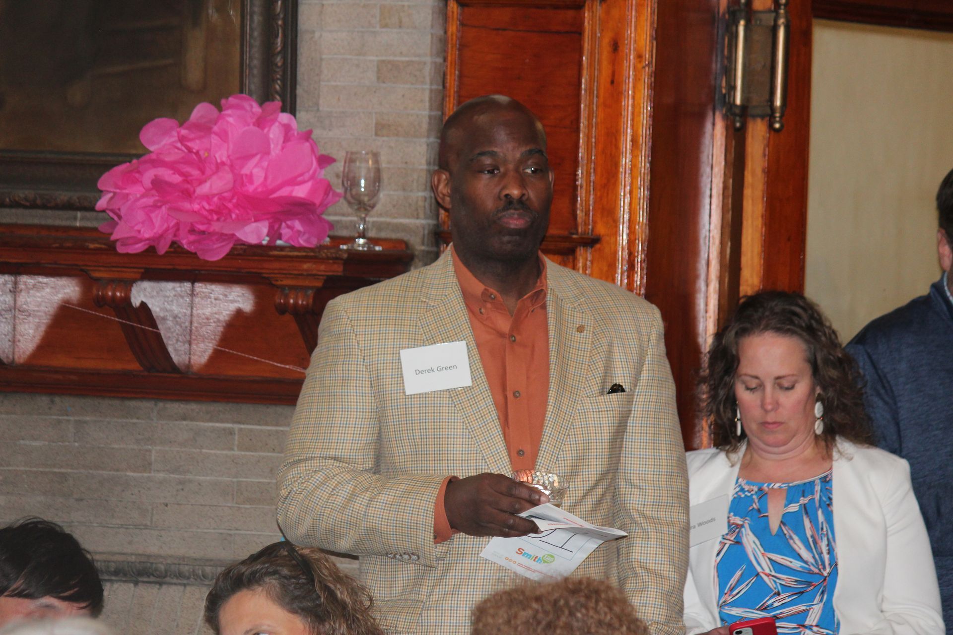 Man in tan blazer stands at an indoor event, holding papers, beside a woman in a white jacket.