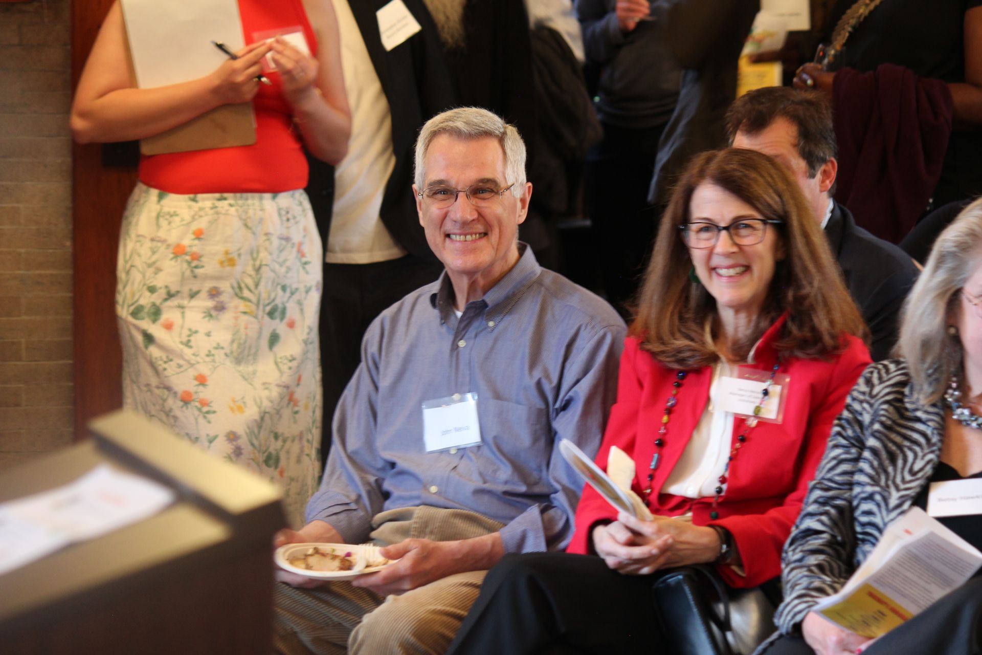 Two smiling people seated in a meeting, holding papers and a plate, with others standing nearby