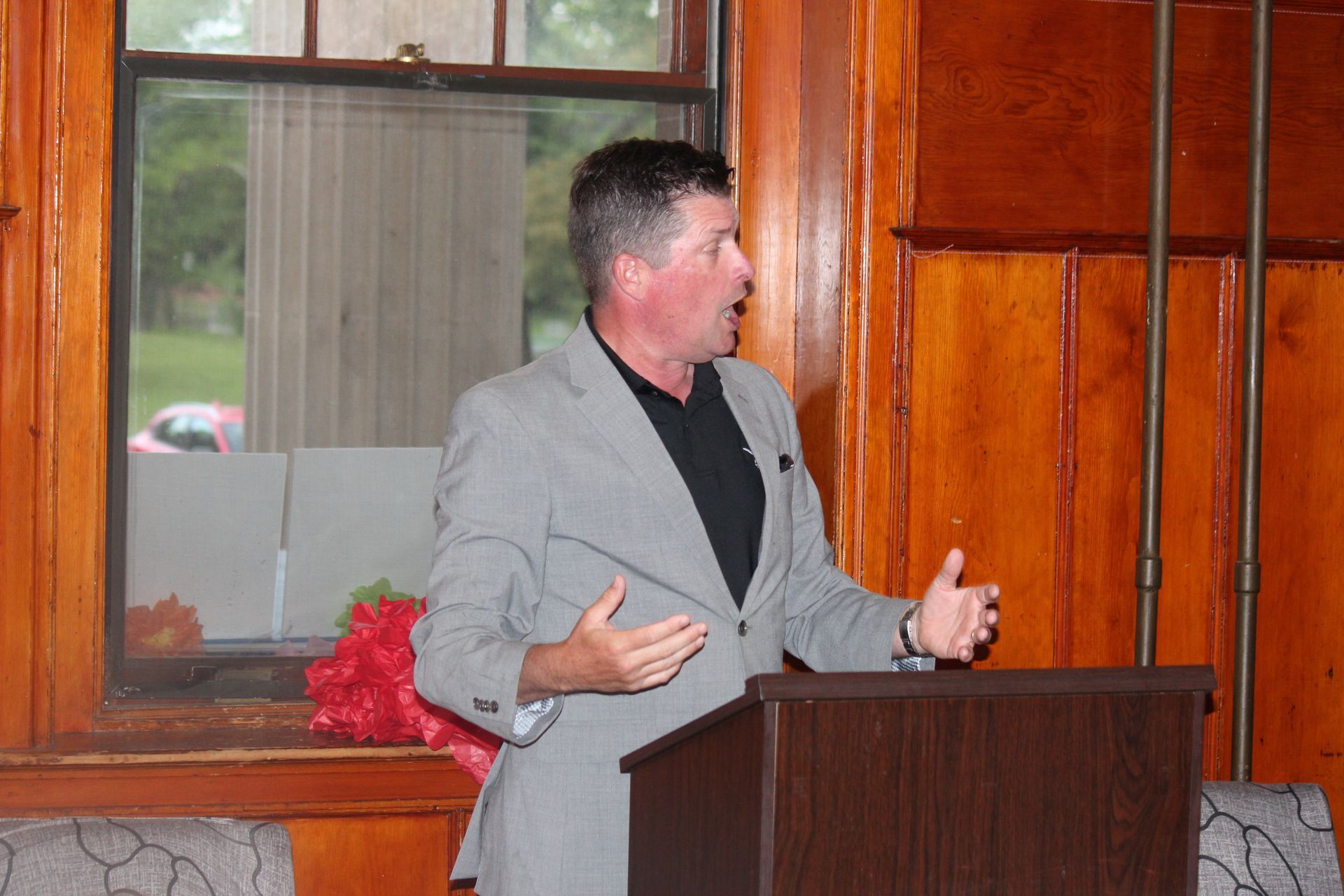 Man speaking at a podium in a wood-paneled room, gesturing with both hands.