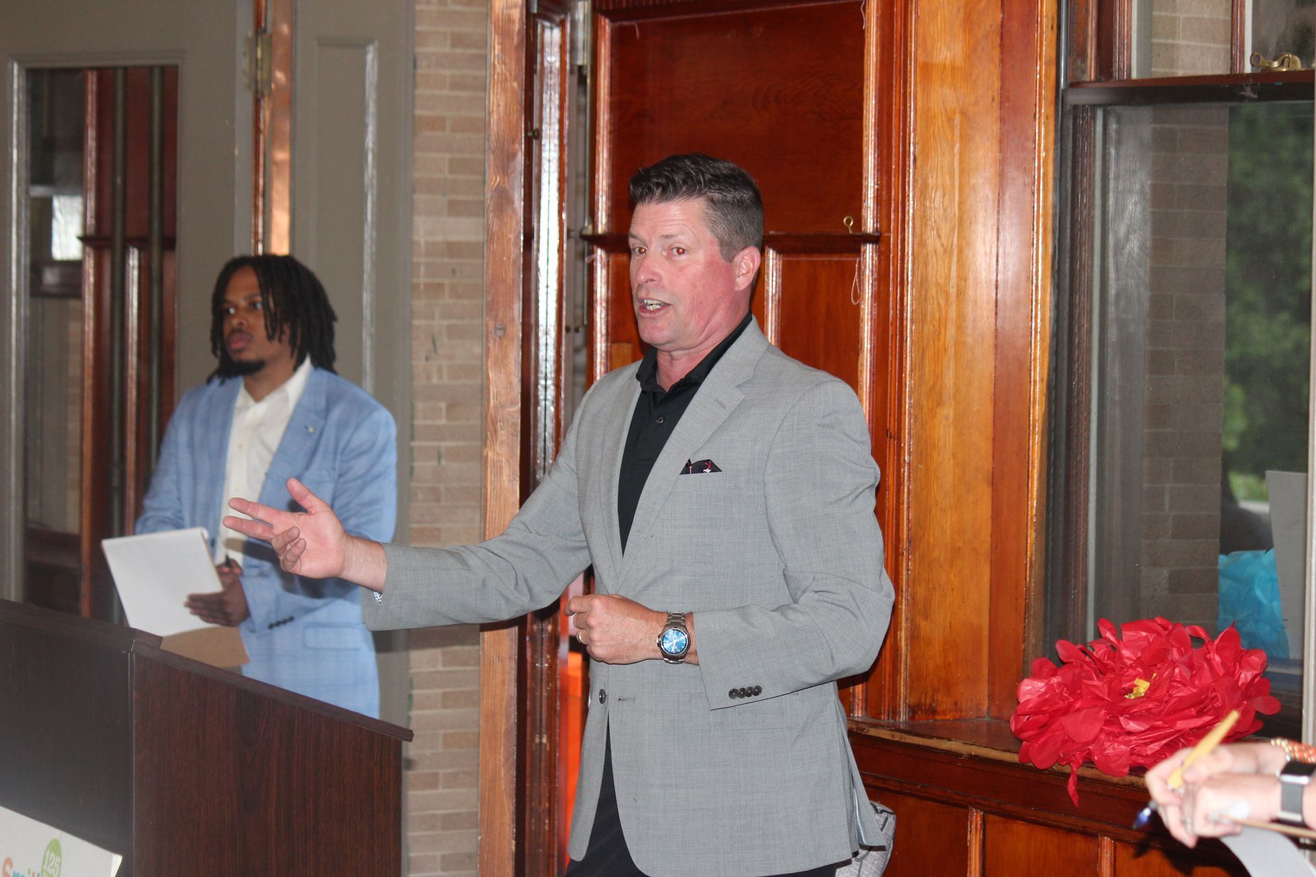 Man speaking at a podium in a wood-paneled room, with another person standing behind him holding papers.