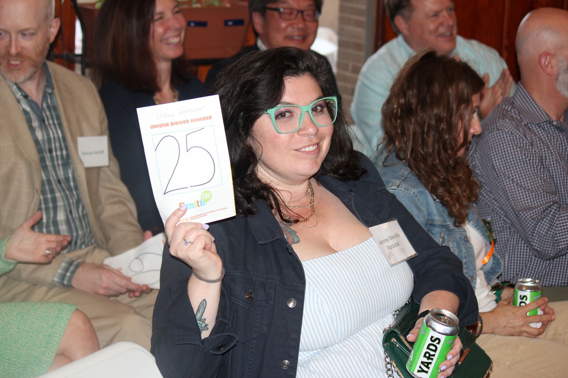 Woman in teal glasses holds up a paper sign and a bottle at an indoor event with seated attendees.