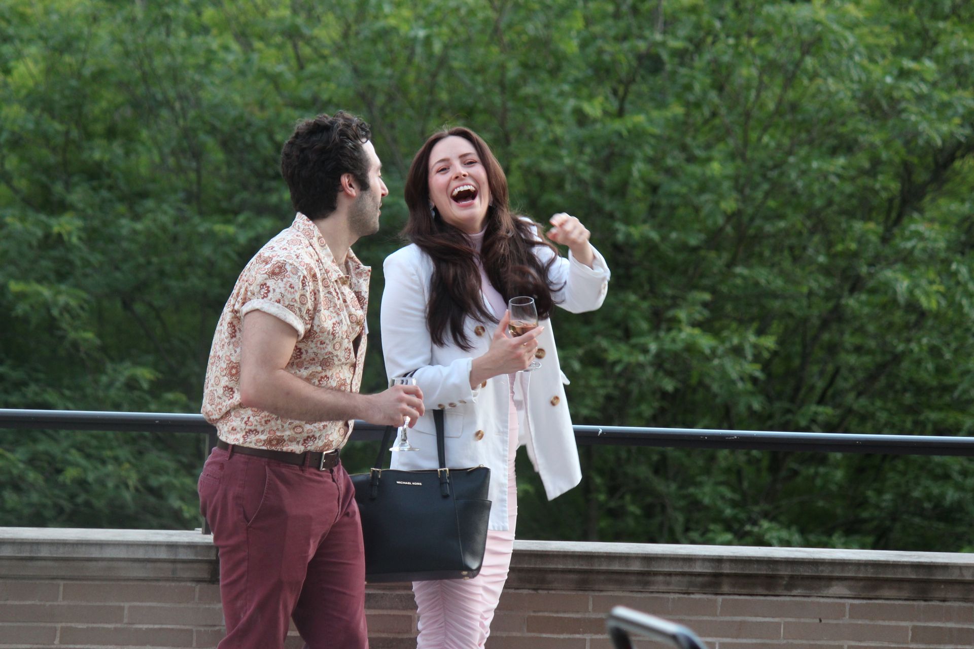 Two people laughing by a railing in a green park, one holding a black purse and coffee cup.