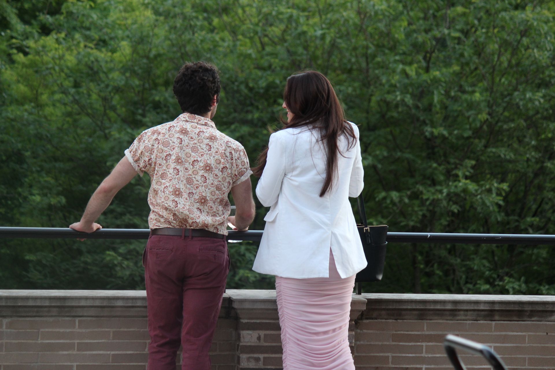 Two people stand at a railing, looking out over green trees.