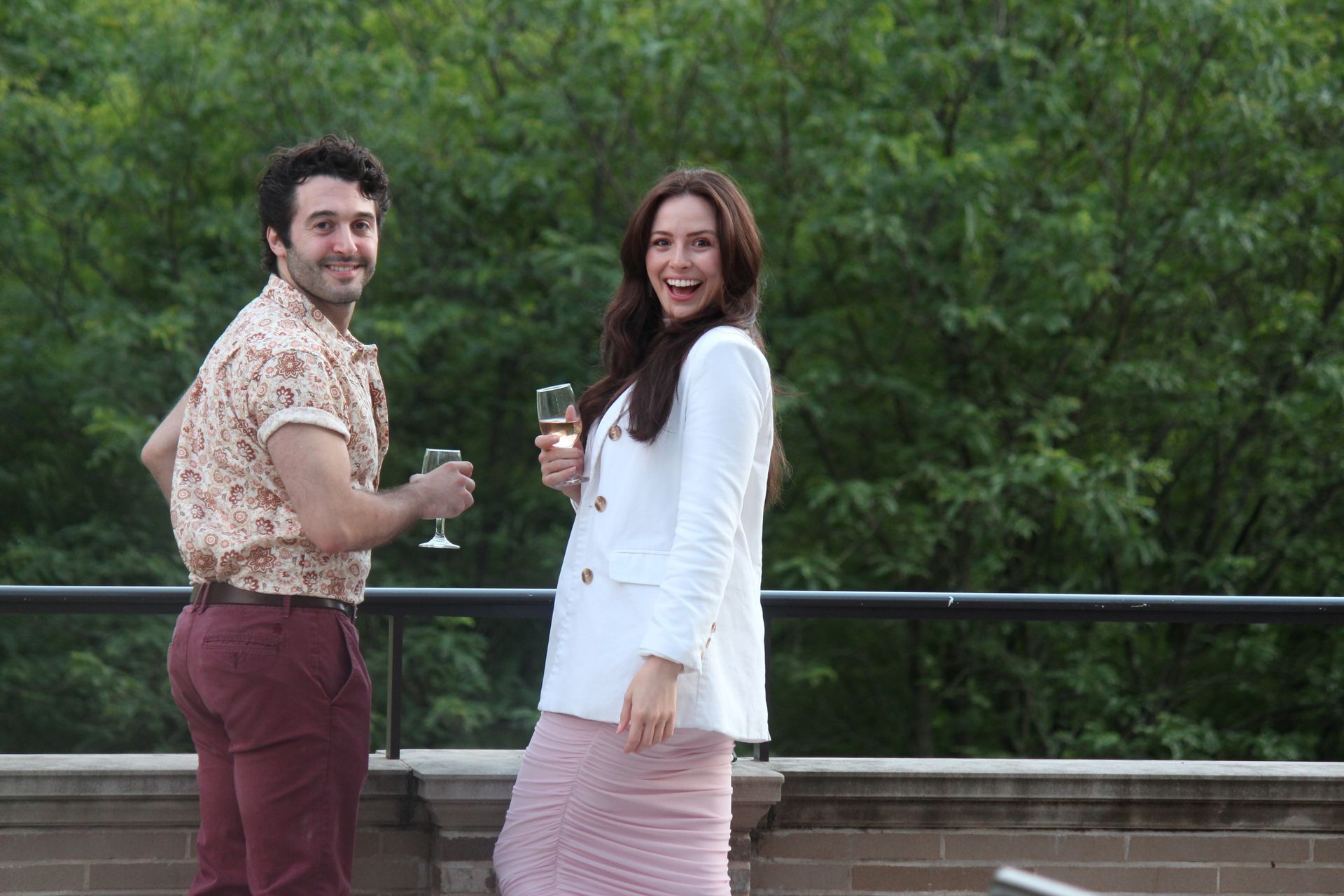 Two people smiling and holding drinks on a balcony with trees in the background