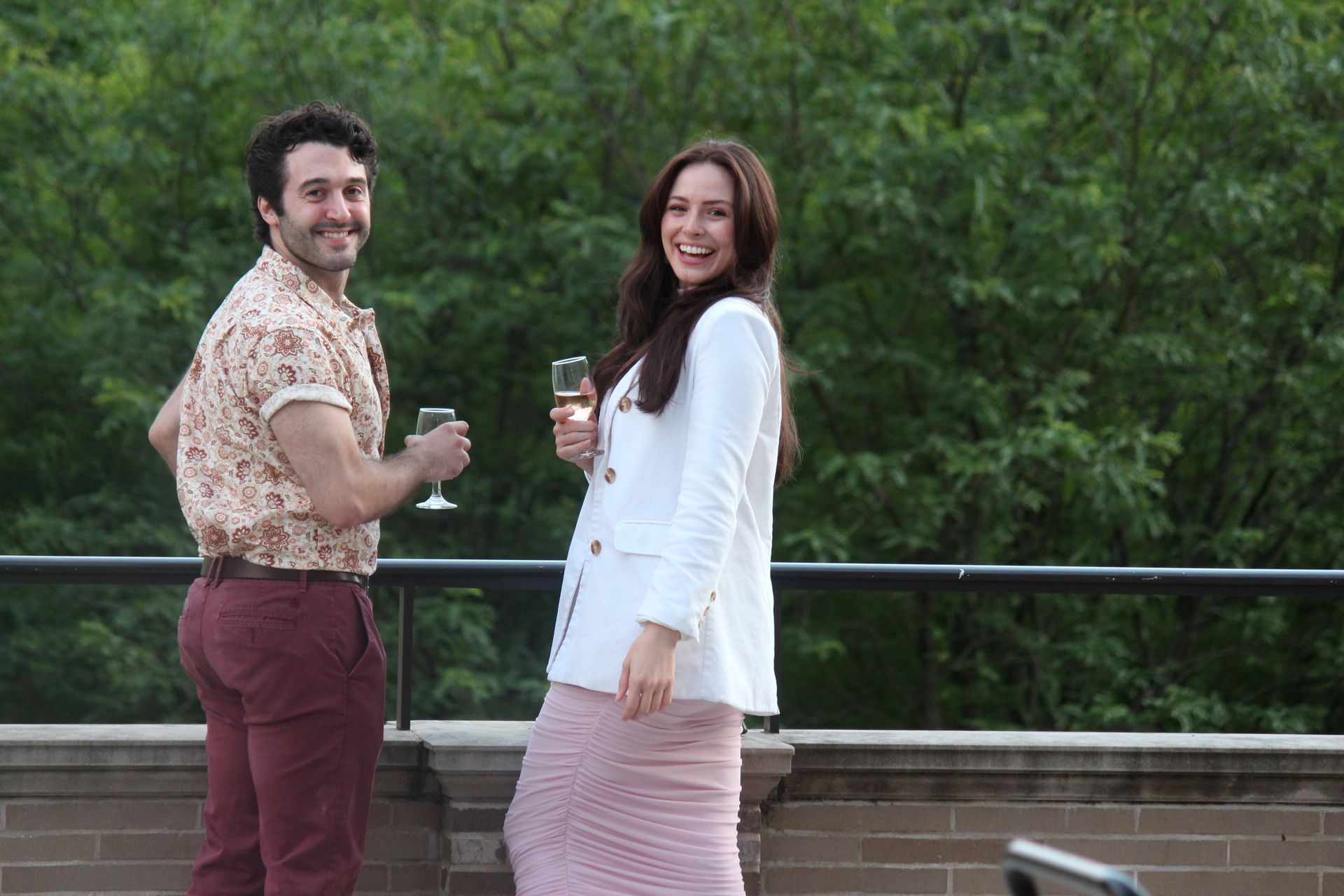 Two people smiling on a terrace, holding drinks, with green trees in the background.