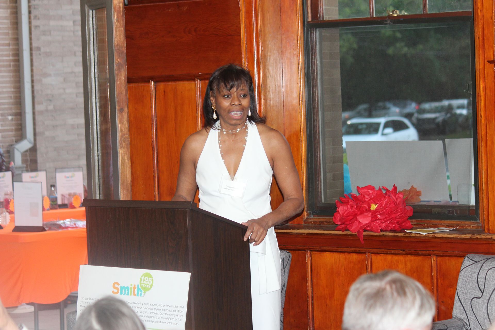 Woman in a white dress speaking at a podium in front of a window and red flowers
