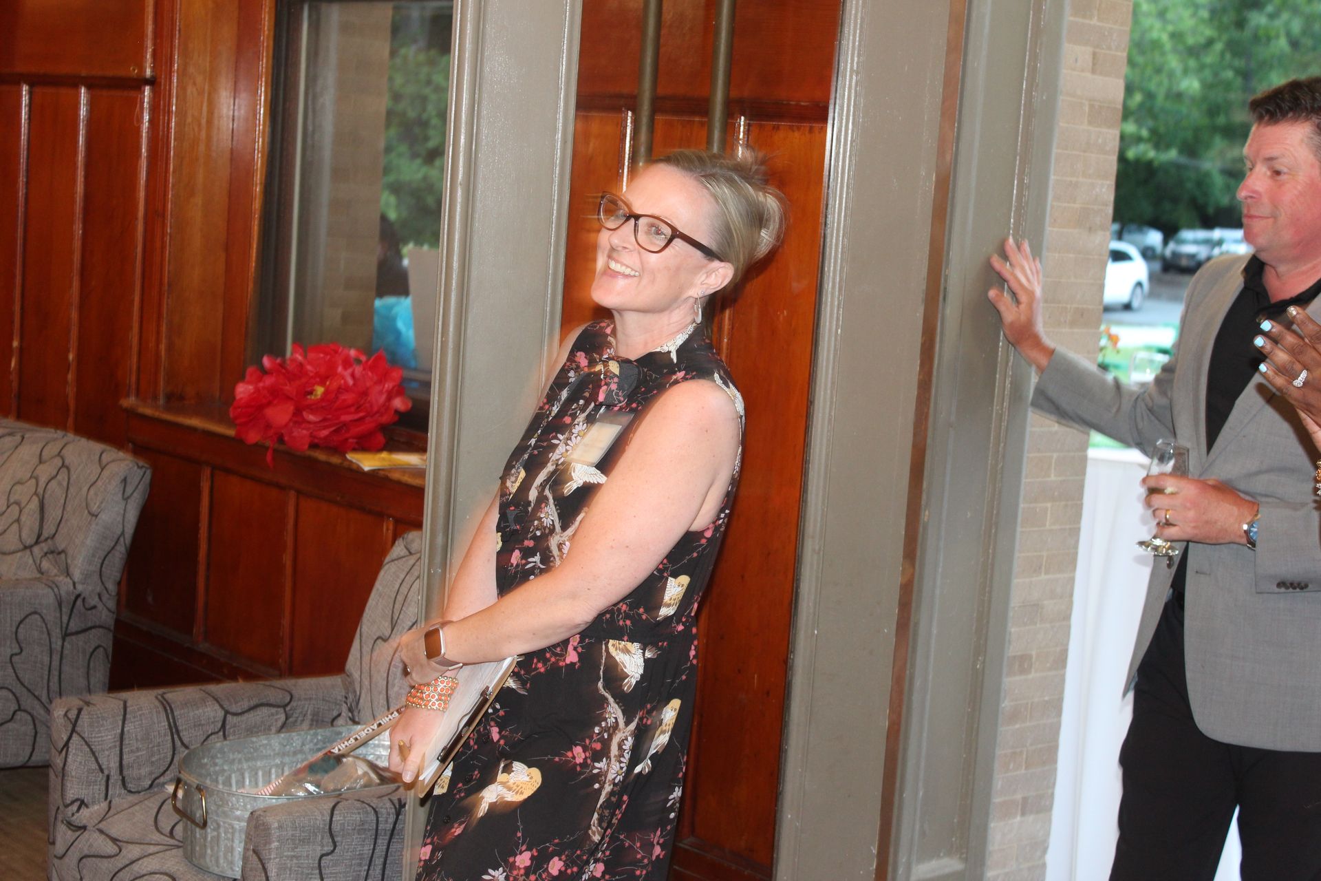 Smiling woman in a floral dress stands indoors near wood paneling, with a man in a blazer behind her.