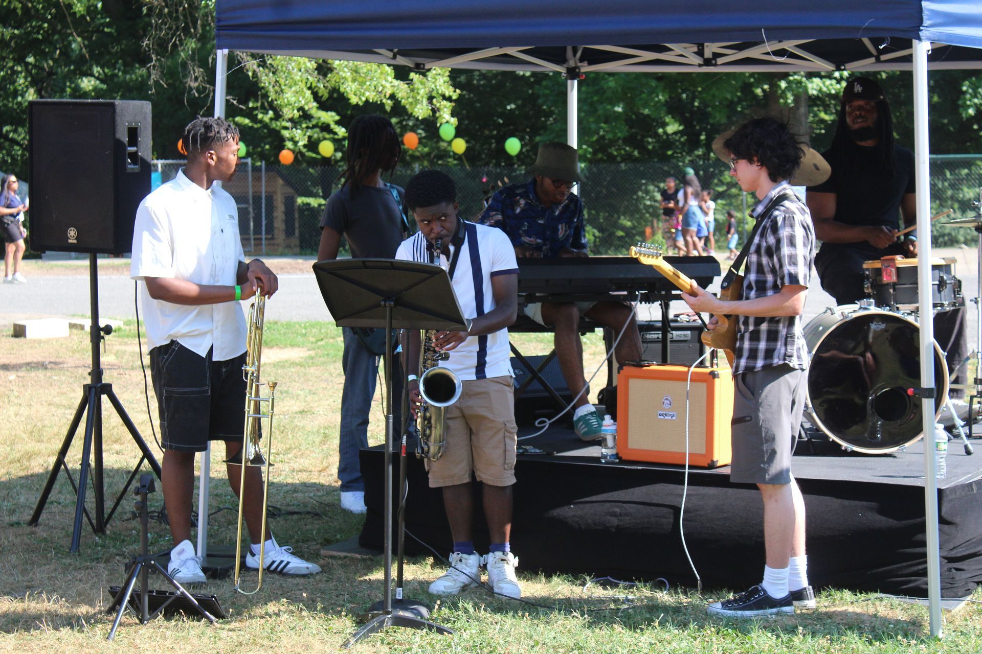 Three musicians perform outdoors under a blue canopy, with saxophone, guitar, and drum kit.
