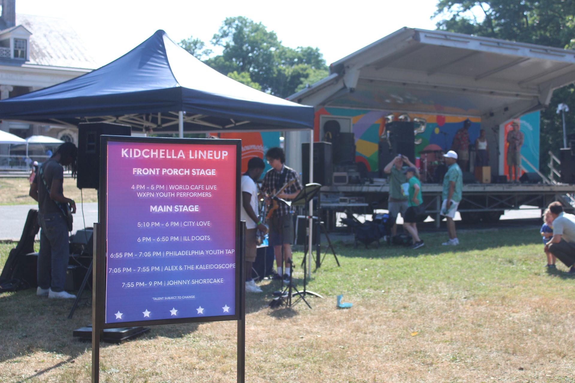 Outdoor festival stage with a purple sign, blue tents, and performers on a sunny grassy field