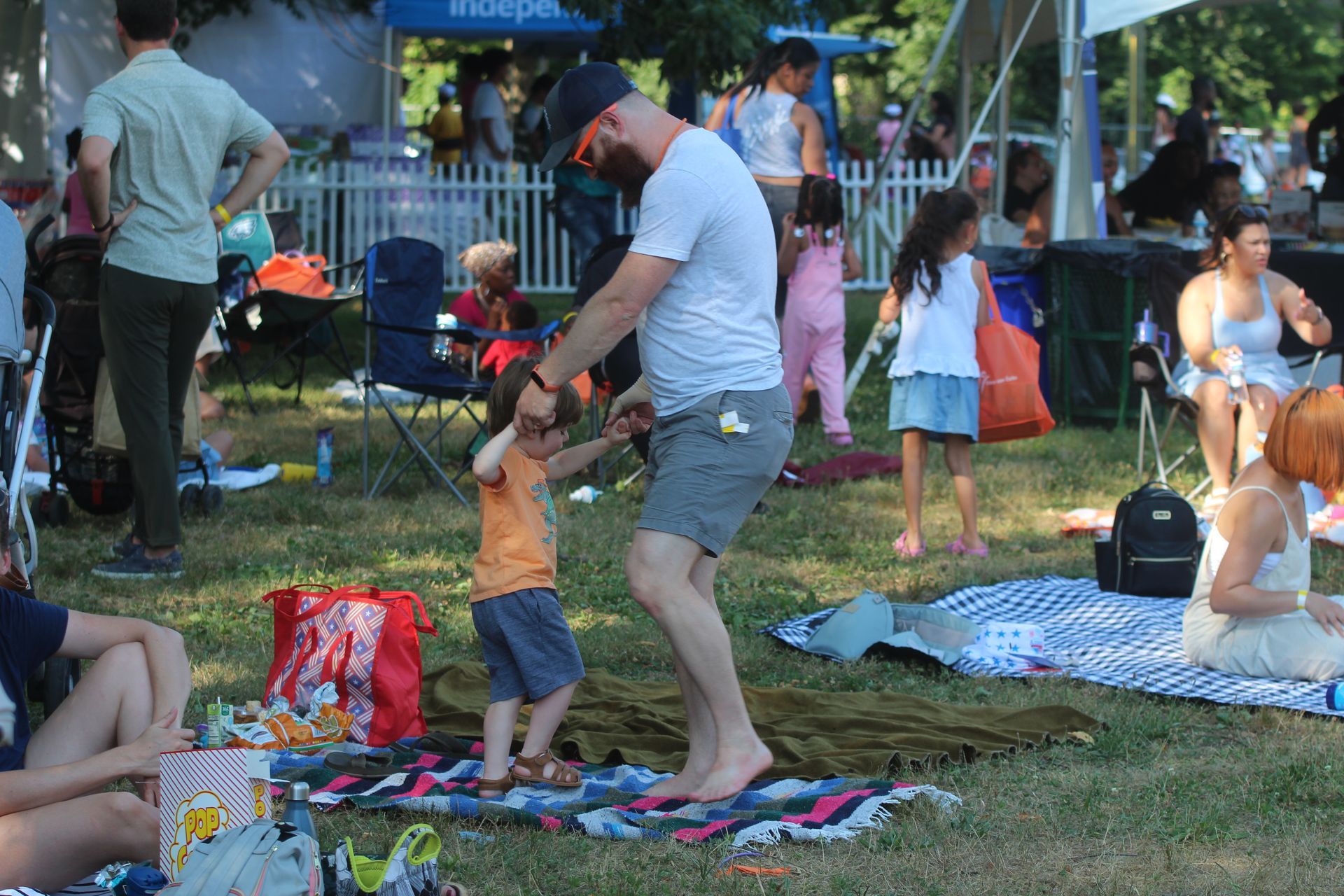 Adults and child on a picnic blanket in a park, with drinks, bags, and people relaxing nearby