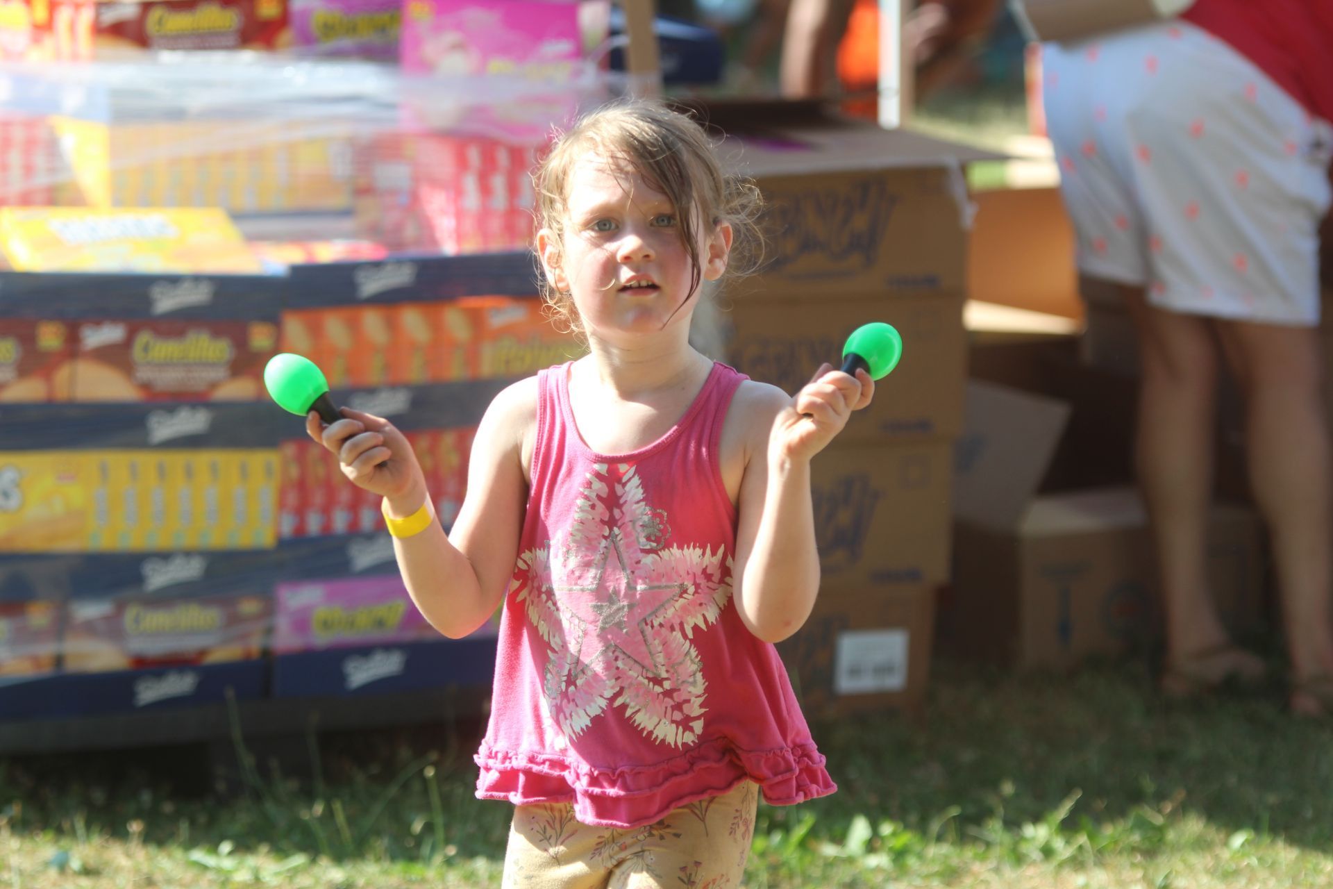 Child in pink shirt holding two green maracas at an outdoor fair.