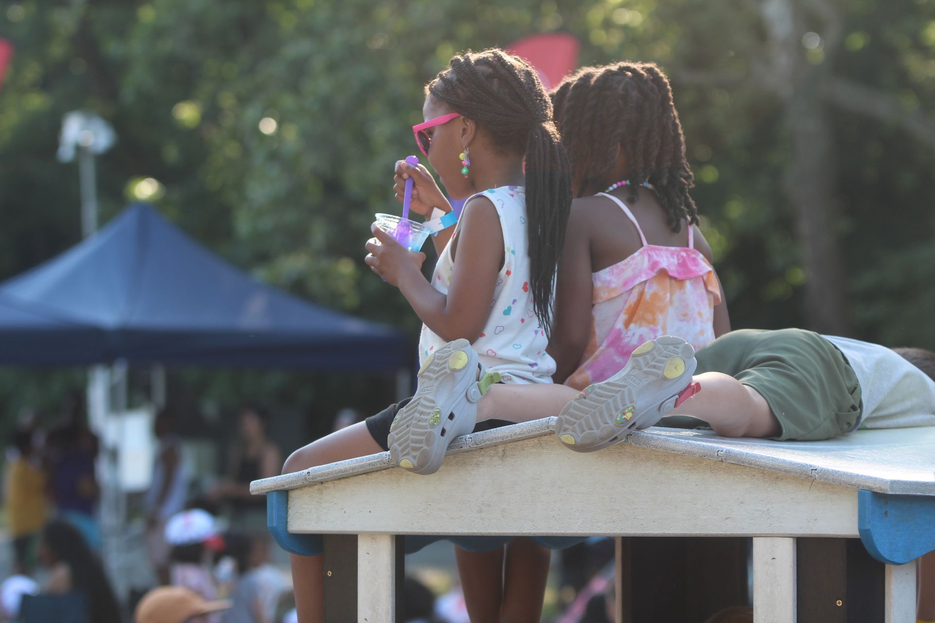 Two children sitting on a platform, one drinking from a straw at an outdoor event