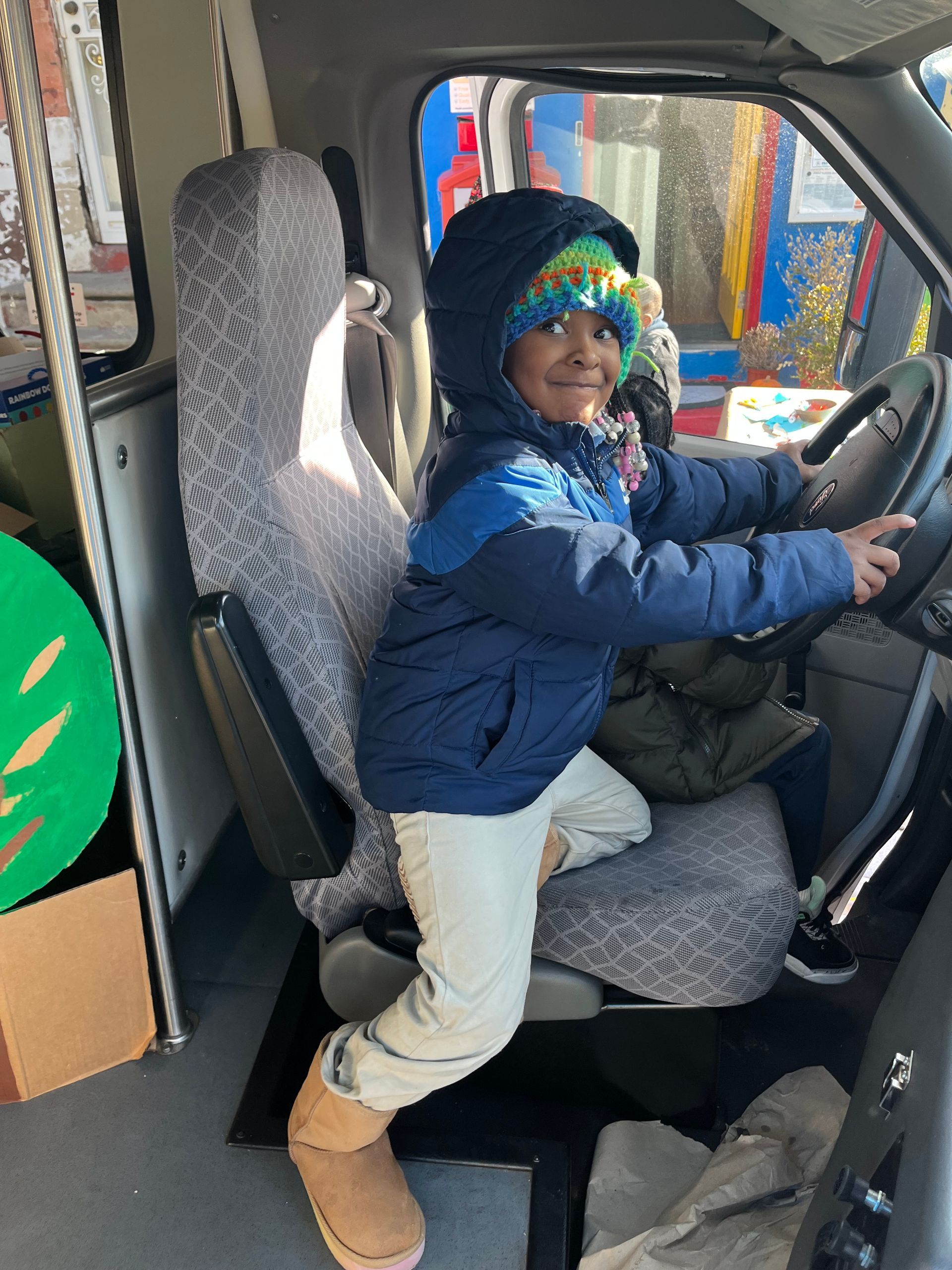 Child smiling while sitting in the driver’s seat of a vehicle, wearing a blue jacket and tan boots.