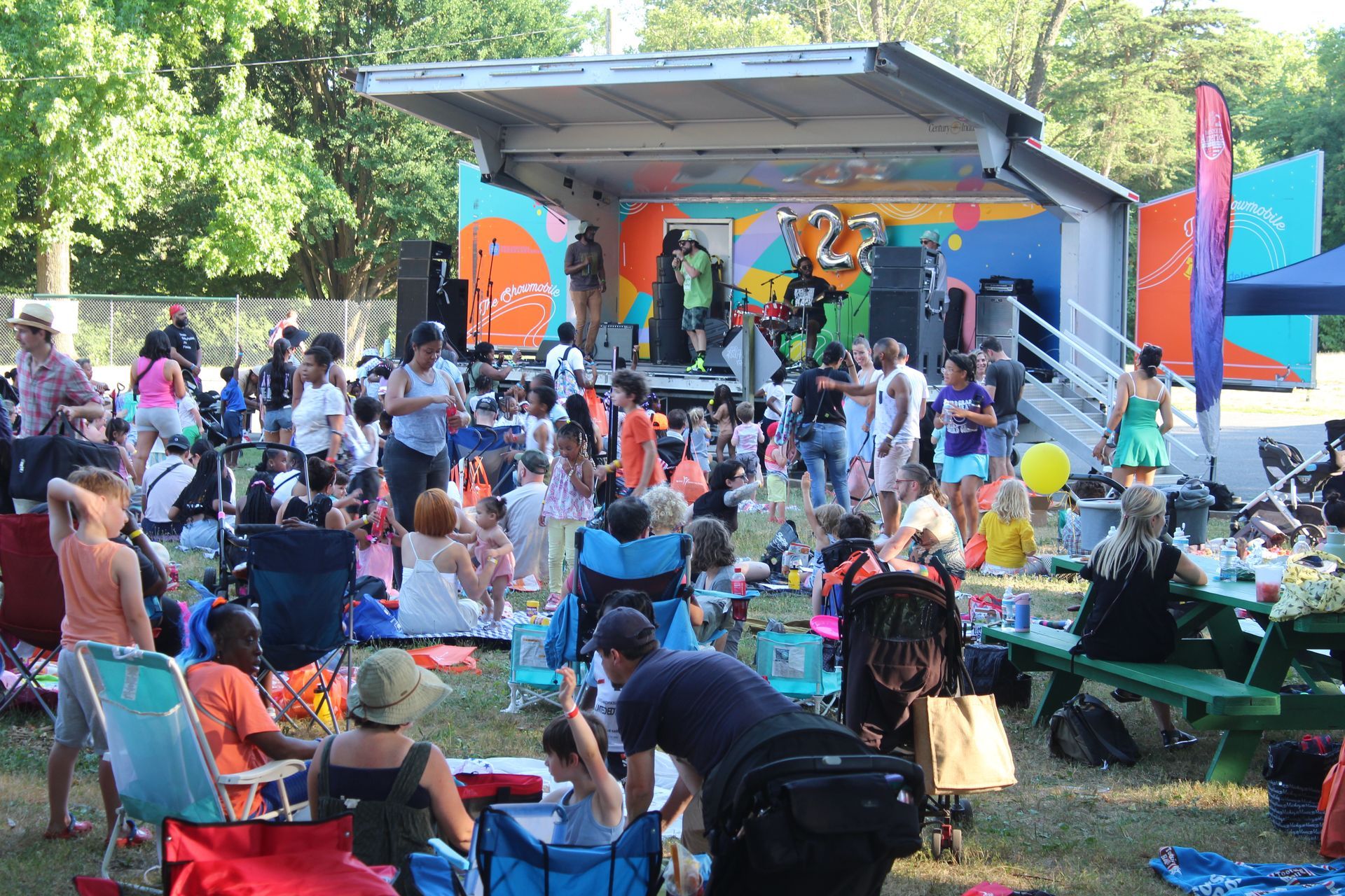 Outdoor concert with a crowd seated on blankets and chairs facing a colorful stage in a park