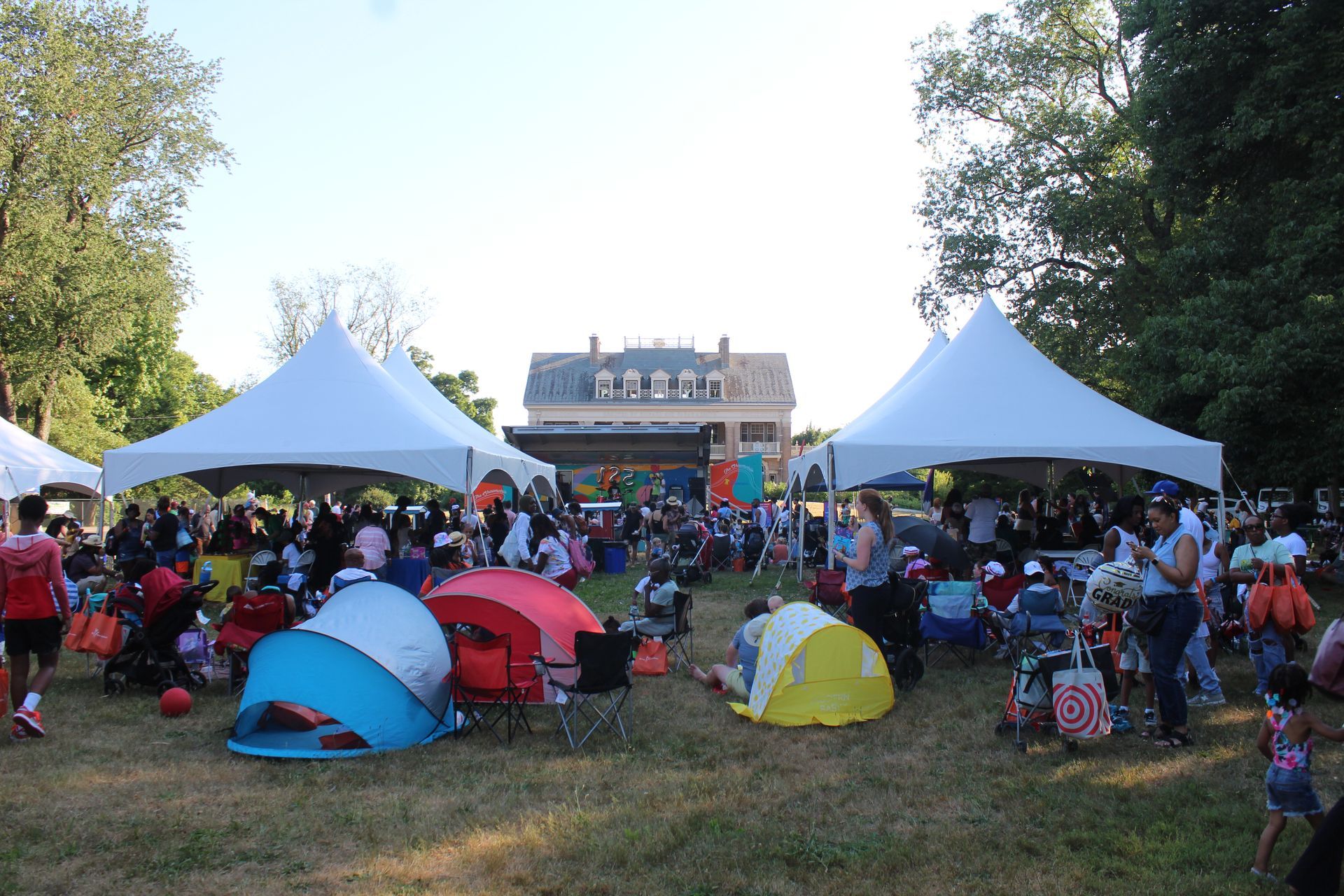 Crowd seated on grass at an outdoor event with white tents and colorful umbrellas under trees
