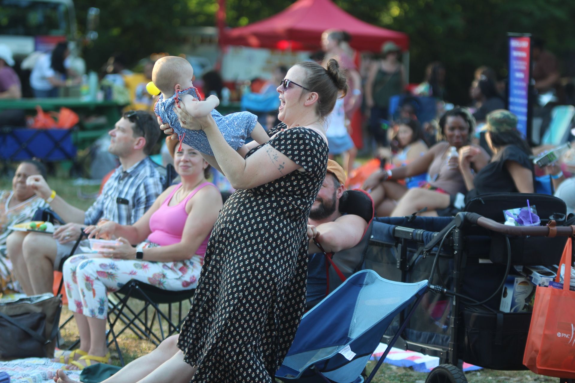 Woman in a black polka-dot dress holds a baby at an outdoor park event with seated spectators nearby