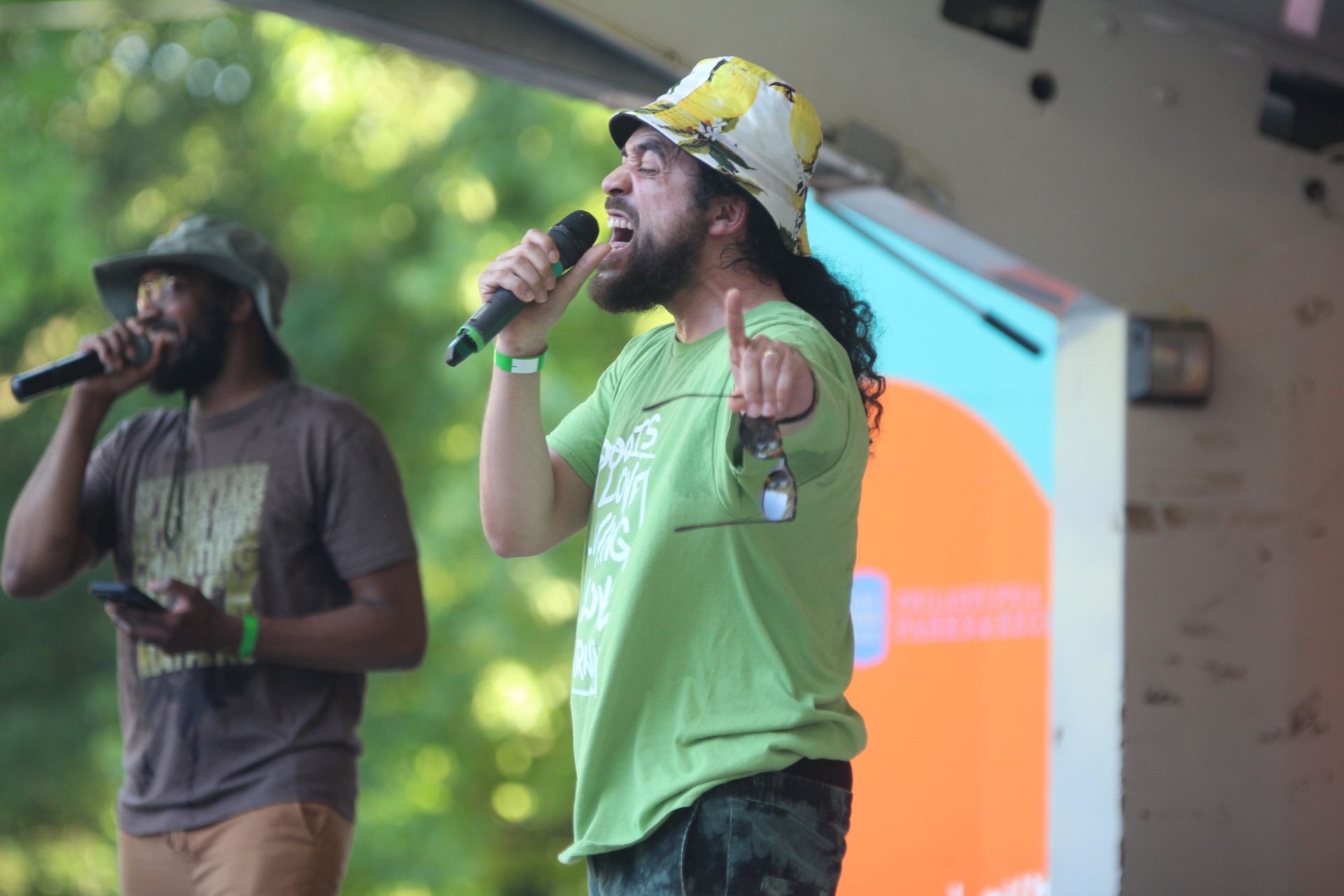 Two singers performing on an outdoor stage, one in a green shirt singing into a microphone.
