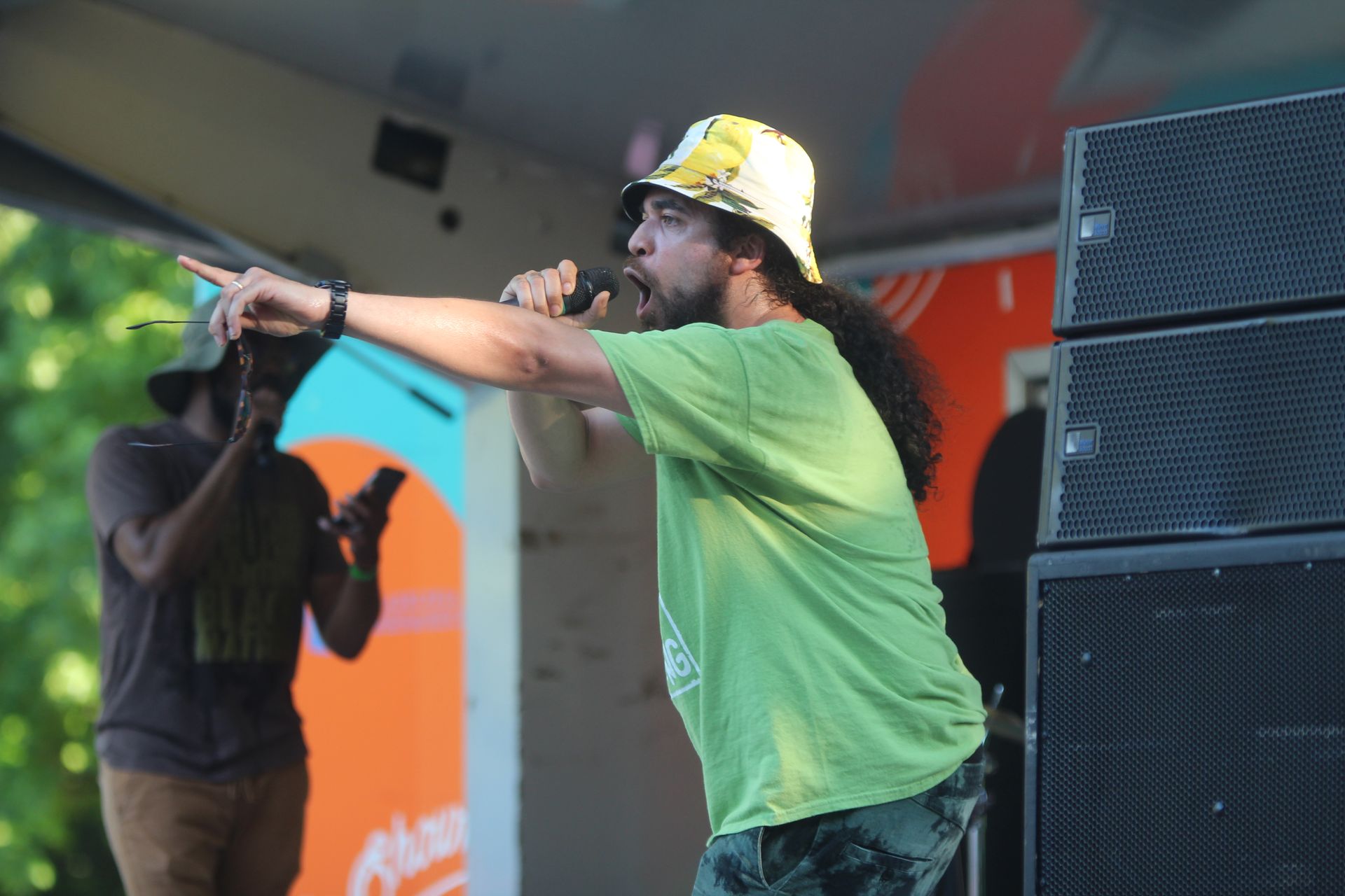 Singer in green shirt and hard hat performing on stage with microphone, beside large speakers