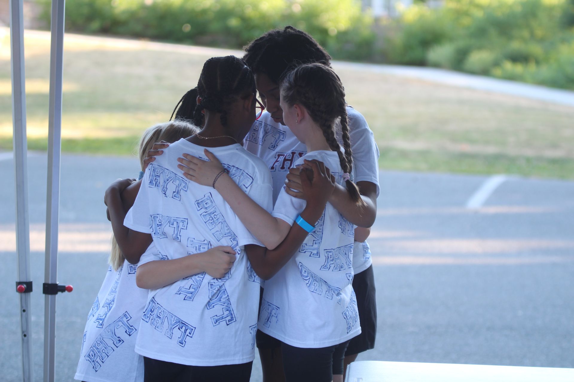 Three people in white shirts hugging together outdoors under a shaded canopy.
