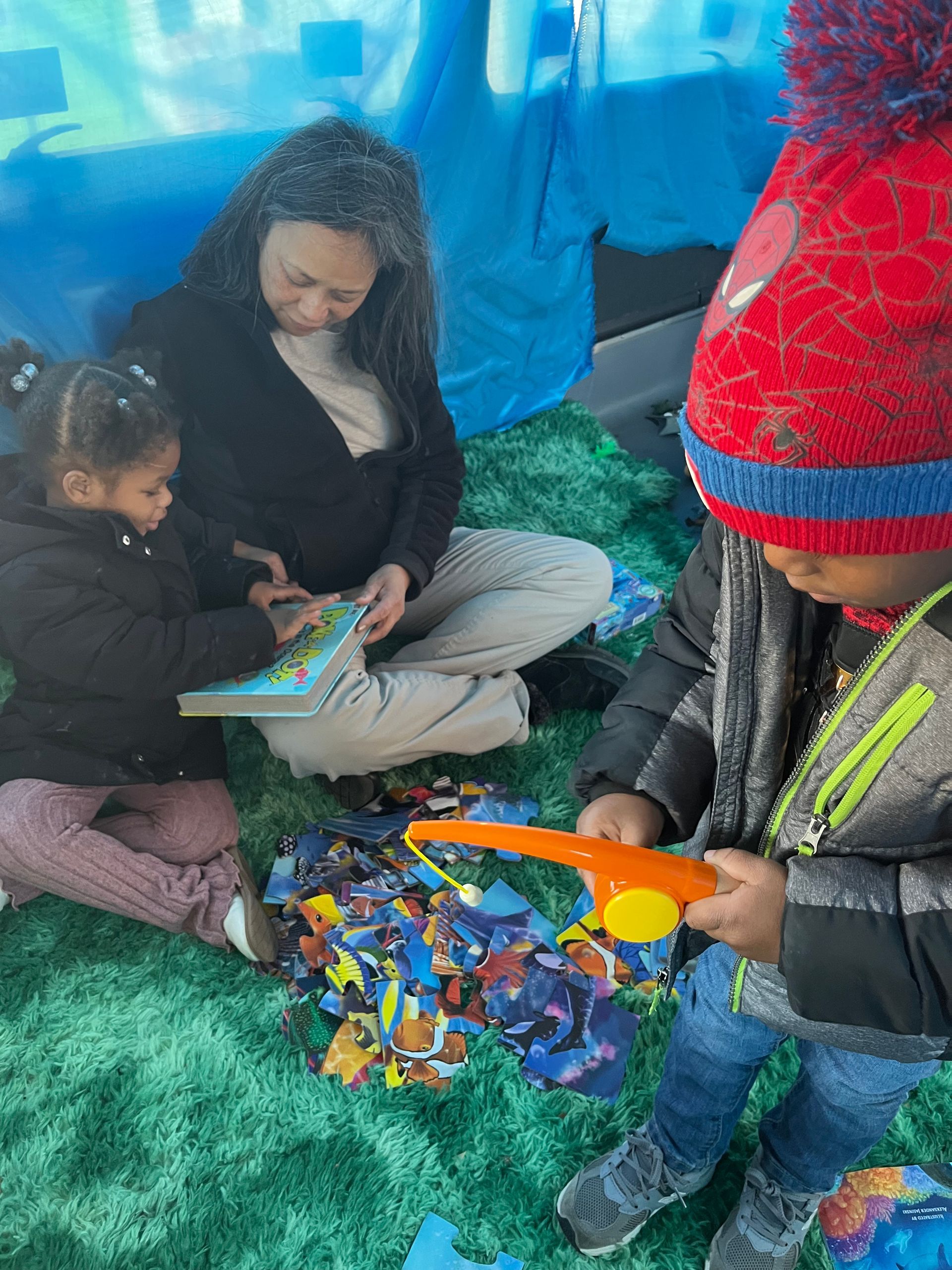 Two children and an adult sitting outdoors, looking at toys in a colorful play area.