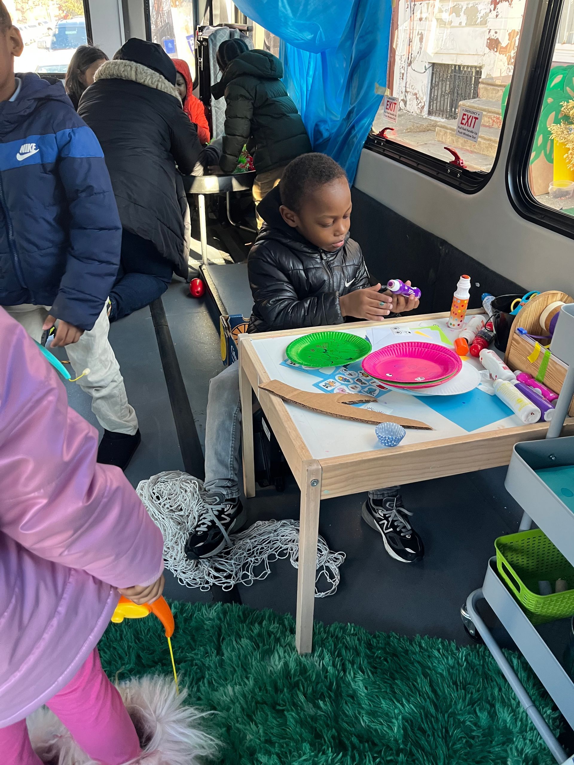 Child painting at a small table inside a bus, with colorful art supplies and a green rug.