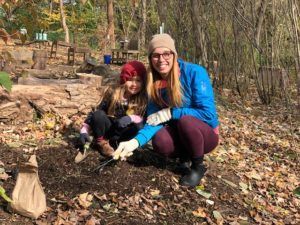Two people crouch outdoors in fall leaves, smiling beside a small dug hole with a sapling.