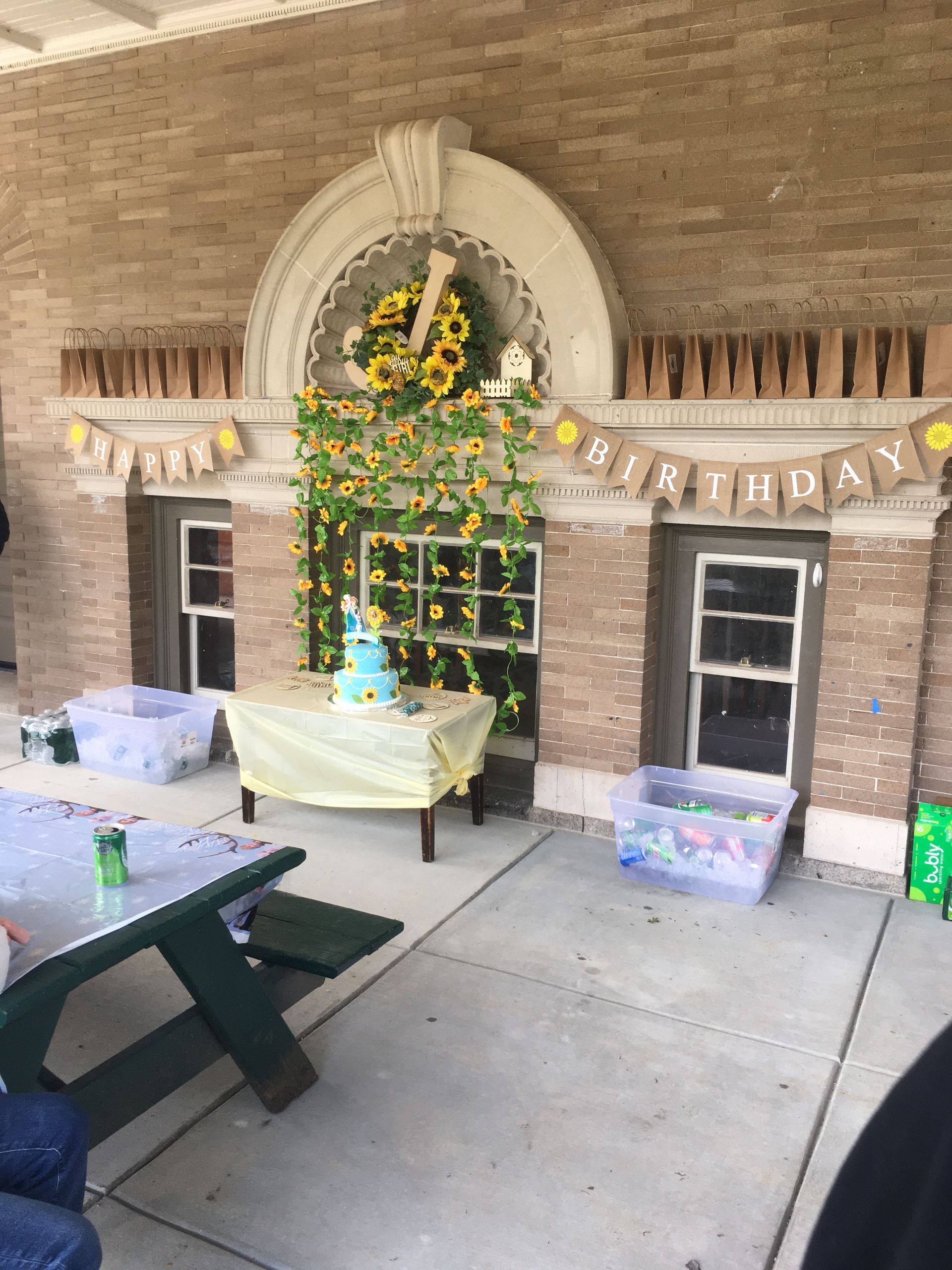 Decorated outdoor table setup under brick archway with yellow flowers and a blue cake at center